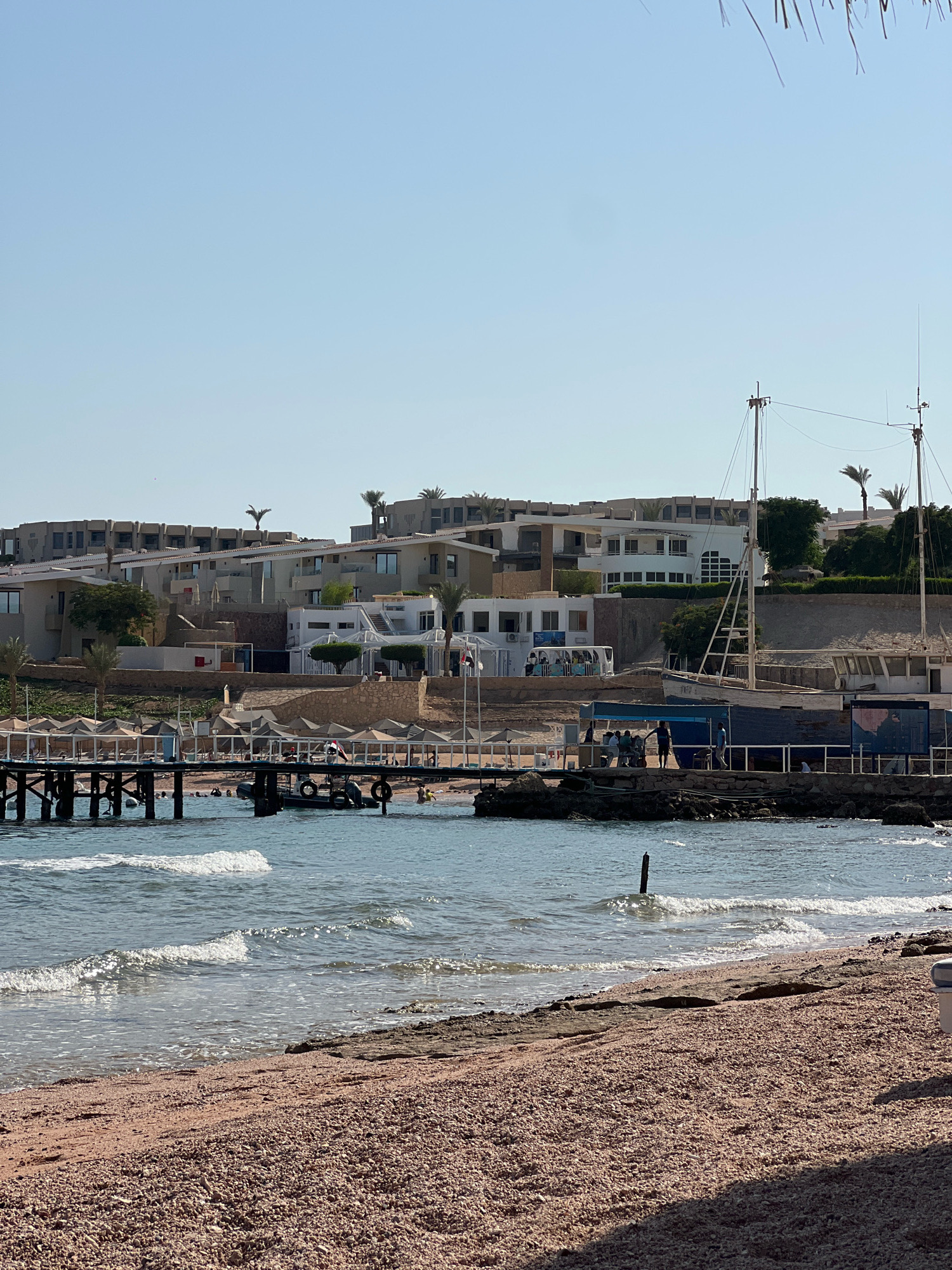The image depicts a coastal scene in Sharm El Sheikh, Egypt, likely at a beach resort. The foreground is a coarse, pinkish-brown pebble beach. Gentle waves lap at the shore, with some white foam visible. In the mid-ground, a wooden pier juts out into the clear blue water. Several beach umbrellas and lounge chairs are set up along the sandy area behind the pier. People are visible in and around the water, some swimming and others standing on the pier. A small inflatable boat with people is tied to the pier.
In the background, a multi-story resort complex is built into a hillside. The buildings are modern with white and beige facades, flat roofs, and large windows. Palm trees and other sparse vegetation are scattered around the resort. Two tall masts, likely part of a boat or for signaling, rise above the buildings on the right side of the image. The sky is a clear, pale blue, indicating a sunny day. The overall impression is one of a relaxed vacation setting by the sea.