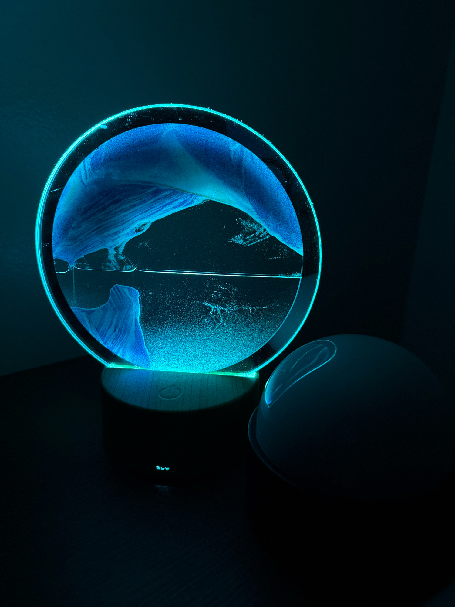 The image displays a circular moving sand art lamp, illuminated by a vibrant teal light, situated on a dark surface. The sand within the lamp is a deep blue, creating abstract patterns that shift and change. The lamp is powered by a dark circular base with illuminated digital numbers on its front. To the right of the lamp, a rounded, dark object, possibly a portable speaker or another decorative item, is partially visible. The background is dark and out of focus, suggesting a dimly lit room or environment. There is no indication of the city of Glastonbury, United States of America.
