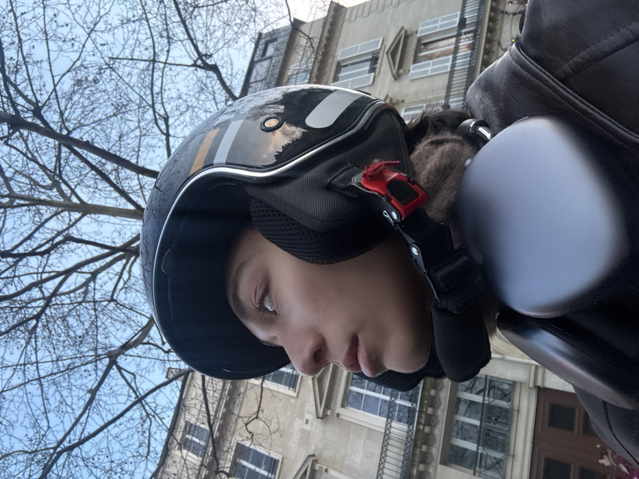 A low-angle, close-up image depicts the right profile of a young individual, appearing female, wearing a black open-face motorcycle helmet adorned with silver and gold-yellow reflective stripes. Her light-colored eyes are directed upwards and to the left. A red quick-release buckle on the helmet's chin strap is visible. Silver-grey over-ear headphones are worn around the neck, resting over a dark brown leather jacket.
The background features a multi-story building constructed of light-colored stone, exhibiting classic Haussmannian architectural characteristics typical of Paris, France. The building includes multiple windows, some with black wrought-iron balconies and white louvered shutters. Above the building, intricate bare branches of a deciduous tree are silhouetted against a light blue sky with minimal cloud cover. The scene is illuminated by natural daylight.