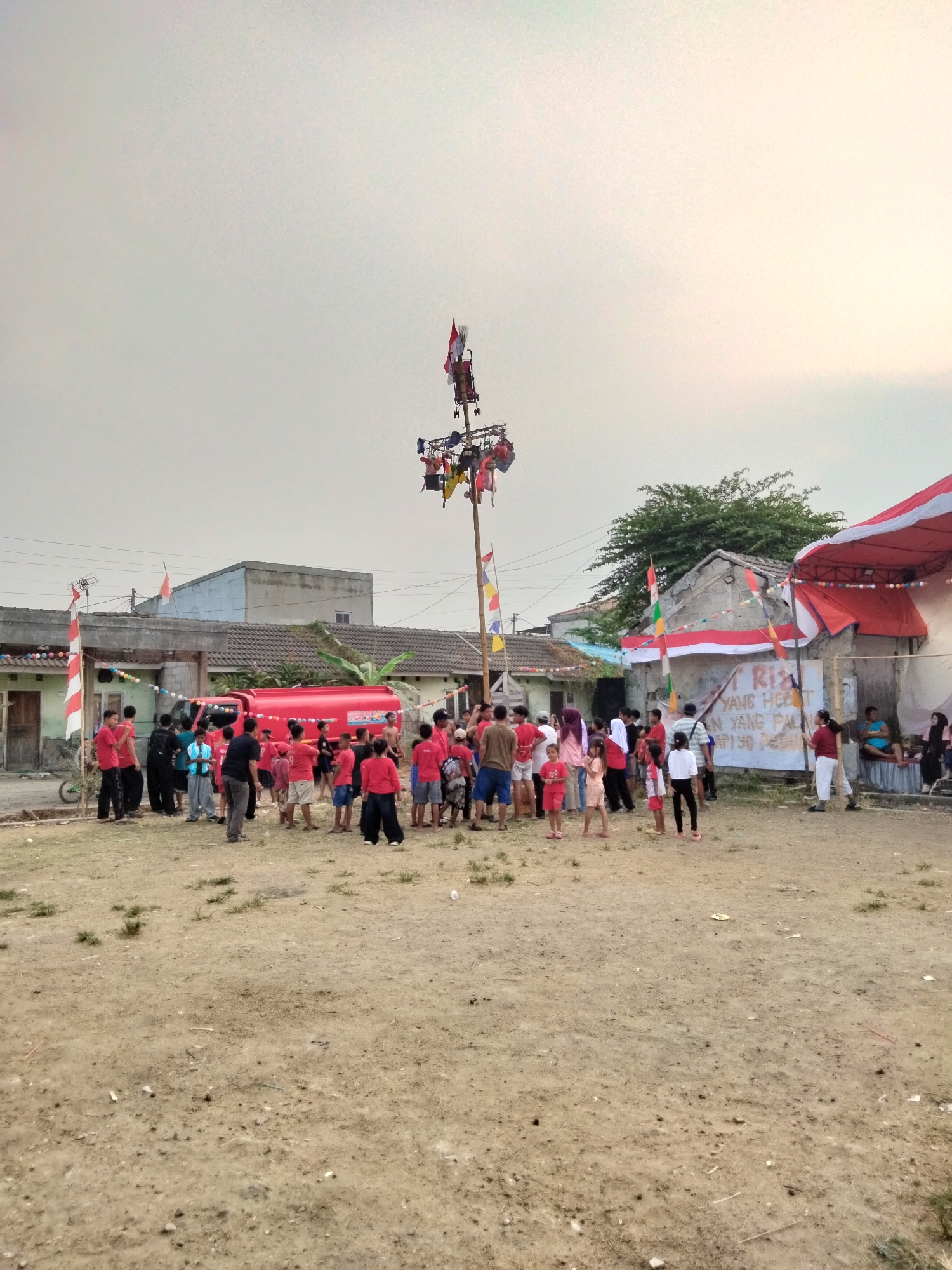 The image depicts an outdoor gathering of people in what appears to be a rural or suburban area in Cariu, Indonesia, likely during a festive occasion. In the foreground, a patch of dry, dusty ground is visible, with sparse tufts of grass and scattered debris. A tall, multi-tiered pole structure adorned with flags and possibly small figures or decorations stands prominently in the center of the scene. Below this structure, a crowd of people, predominantly children and young adults, are gathered. Many are wearing red clothing, suggesting a possible theme or uniform for the event. To the left, a bright red truck, possibly a fire truck given its color and design, is parked.
Behind the crowd, several simple residential buildings with tiled roofs are visible, some appearing slightly weathered. Red and white Indonesian flags are displayed on some of the buildings and on poles. A temporary red and white tent or canopy structure is set up on the right side of the image, where a few individuals are seated. A banner with text in Indonesian is affixed to a wall or fence behind the tent. The overall atmosphere appears to be one of community celebration, possibly related to a local holiday or event. The sky is overcast, indicating a diffused light condition.