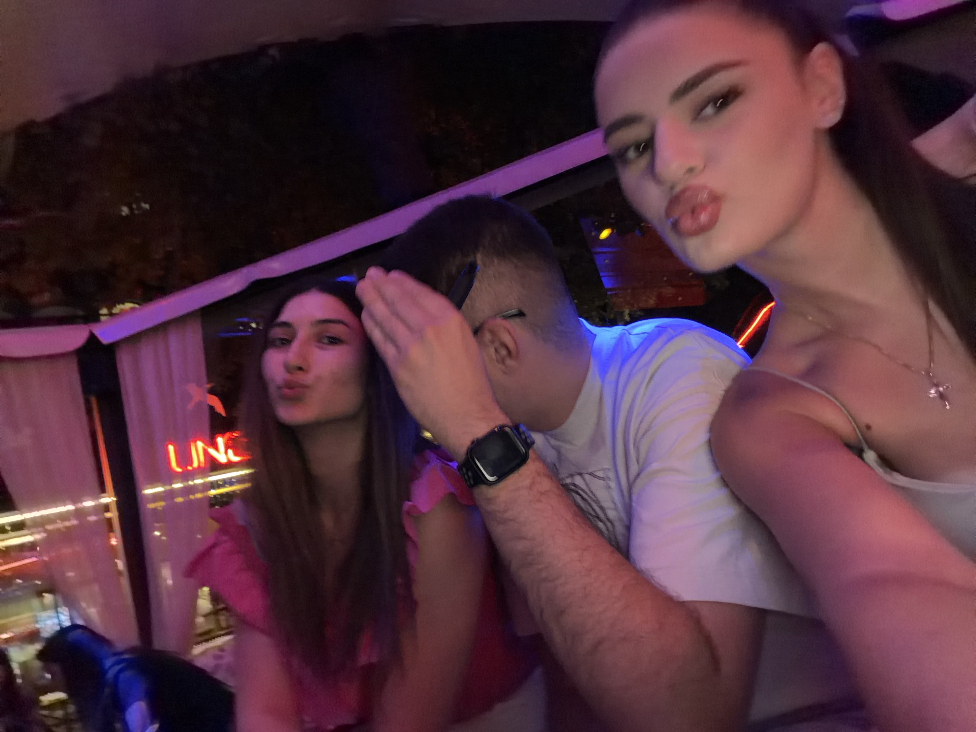 Three individuals, two women and one man, are captured in a close-up, selfie-style photograph. They are situated in an indoor setting, possibly a restaurant or bar, with soft, ambient lighting dominated by pink and purple hues. In the background, neon lights, including a sign that reads "LUNG", are visible through a sheer curtain, suggesting a nighttime scene in the city of Haskovo, Bulgaria.
The woman on the right, in the foreground, is looking towards the camera with her lips puckered, a gesture often associated with taking a selfie. She is wearing a light-colored top with a delicate necklace featuring a cross pendant. The man in the center is partially obscured, with his face turned away and his hand raised to his forehead, potentially shielding his eyes or adjusting something. He is wearing a white t-shirt and a black smartwatch on his left wrist. To the left, another woman is also looking towards the camera, her lips pursed, and she is wearing a bright pink top. The focus of the image is on the faces and upper bodies of the individuals, creating an intimate and candid feel. The overall atmosphere suggests a social gathering in a lively urban environment.