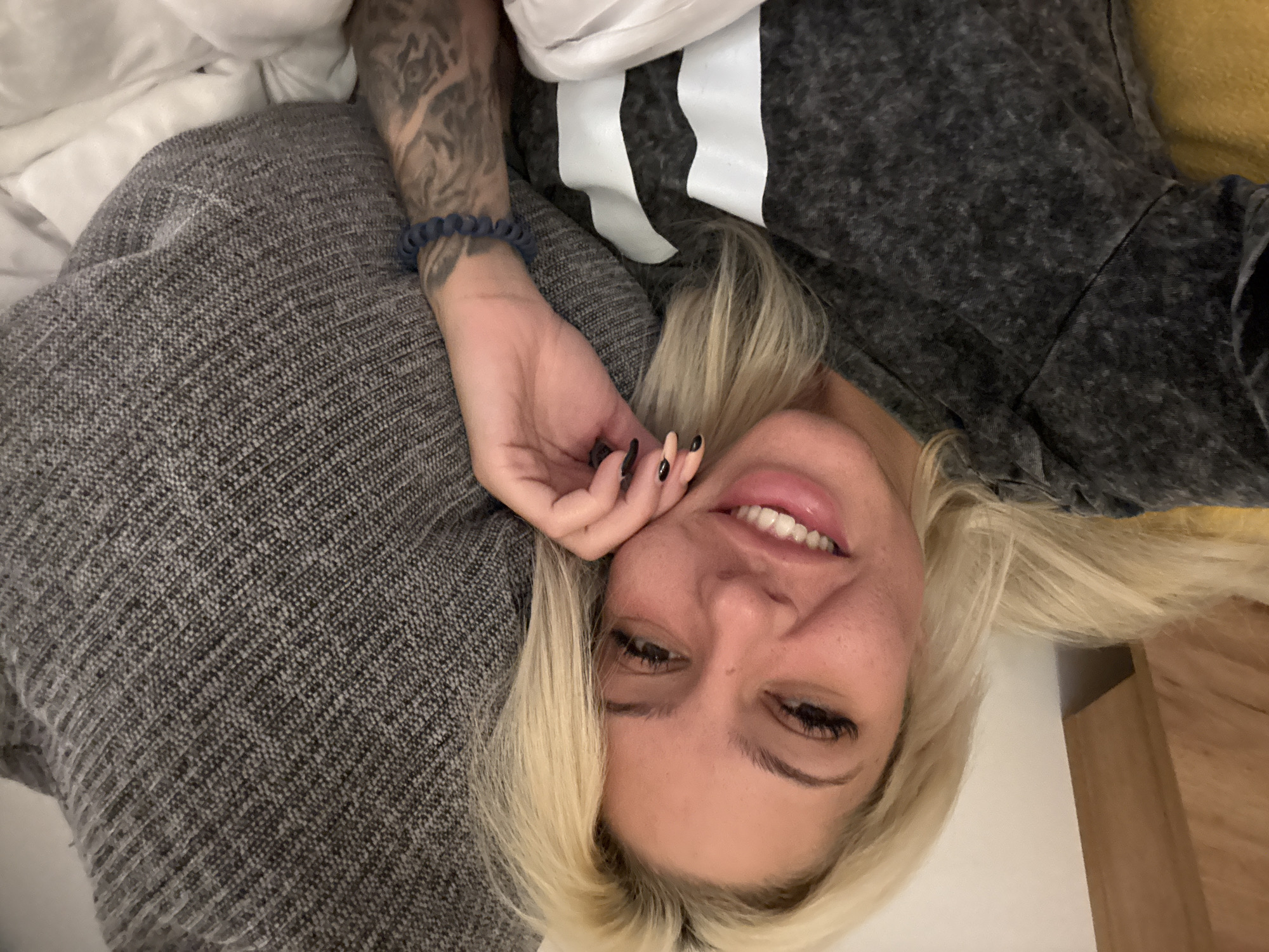 A blonde-haired woman with a tattooed arm and beaded bracelet is lying down, smiling, with her hand near her face. She wears a gray shirt with white stripes. The background includes bedding and a wooden surface. The location is not discernible from the image, and therefore cannot be pinpointed to Sofia, Bulgaria.