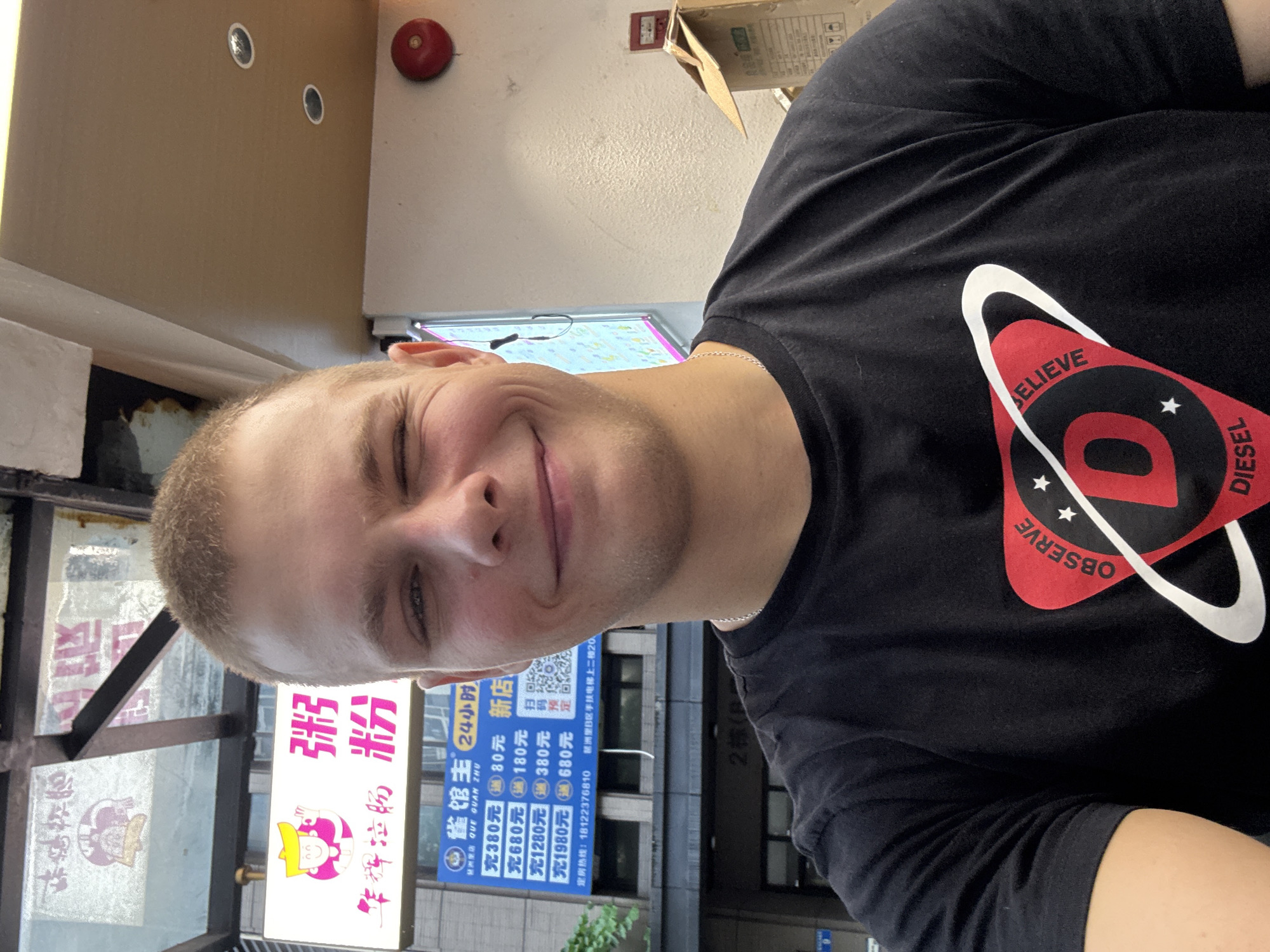 This is a medium shot, eye-level, portrait of a young, fair-skinned man with short blonde hair. He is looking directly at the camera with a slight smile and his eyes are partially closed, giving a playful wink. He is wearing a black t-shirt with a red and white graphic design featuring text that reads "BELIEVE OBSERVE DIESEL". The man is positioned in the foreground, taking up the majority of the frame.
In the background, there are several signs and elements that provide context. To the left, there's a window with various advertisements or signs, one of which appears to be for "华辉拉肠粉" (Hua Hui La Chang Fen). To the right, another sign reads "雀馆主 24小时 新店" (Que Guan Zhu 24 Hours New Store), with pricing information like "充380元 80元" (Charge 380 Yuan Get 80 Yuan) and a phone number "18122376810". Further in the background, a building with the number "2栋(B)" (Building 2 (B)) is visible. The overall setting appears to be an urban street or commercial area, possibly a shop or restaurant, in Haizhu District, China, given the text on the signs. The lighting suggests it is daytime. The man's expression conveys a casual and friendly mood.