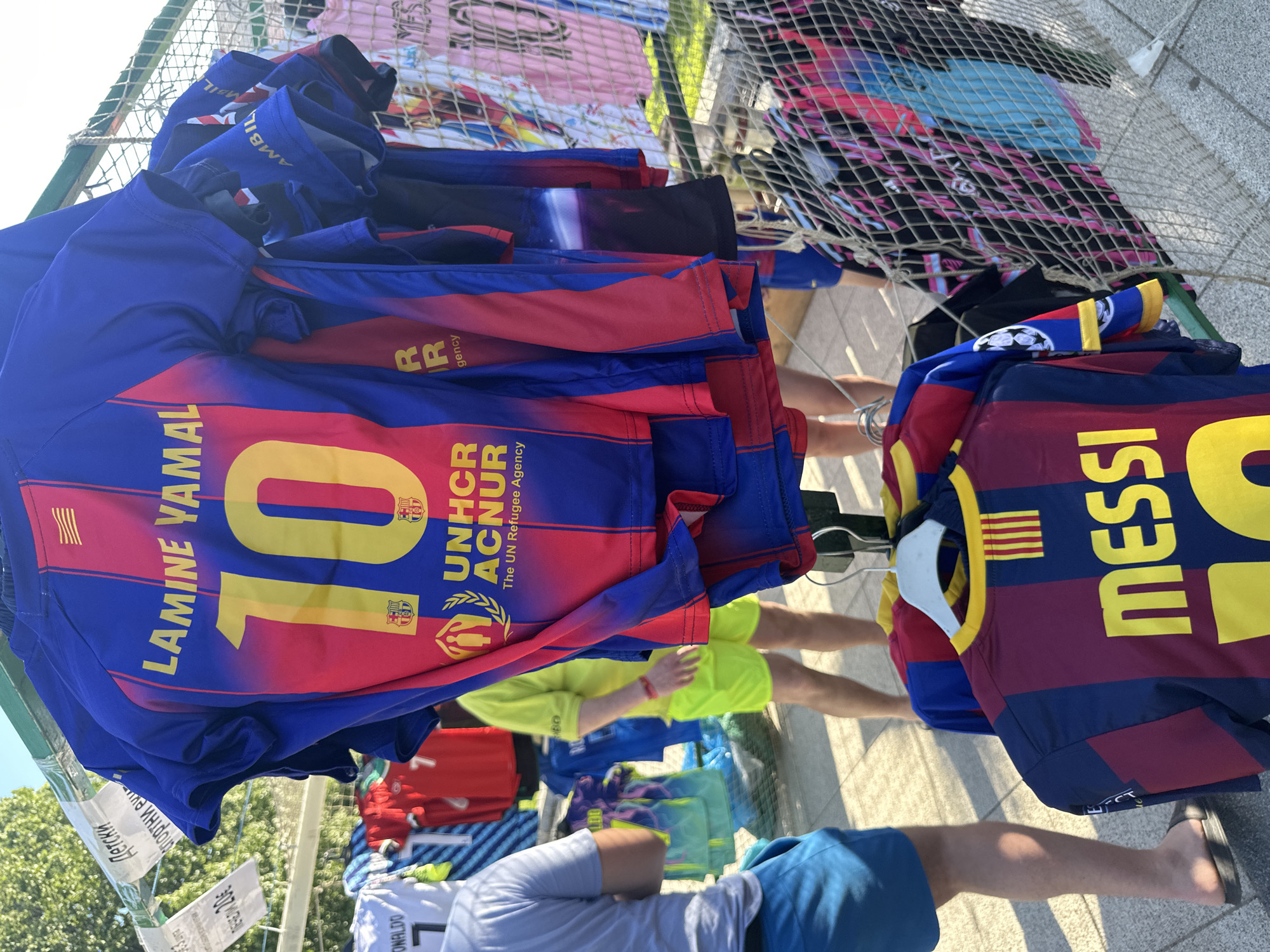 The image shows a rack of soccer jerseys, likely for sale. The jerseys are blue and red striped with yellow lettering. One is labeled "LAMINE YAMAL" with the number "10" and the UNHCR logo. Another is labeled "MESSI". There are also jerseys labeled "RONALDO" hanging on the rack. There is netting in the background. There are several people standing around the rack. The location is likely Montana, Bulgaria, based on the OCR text "Детски Спортни еми".
