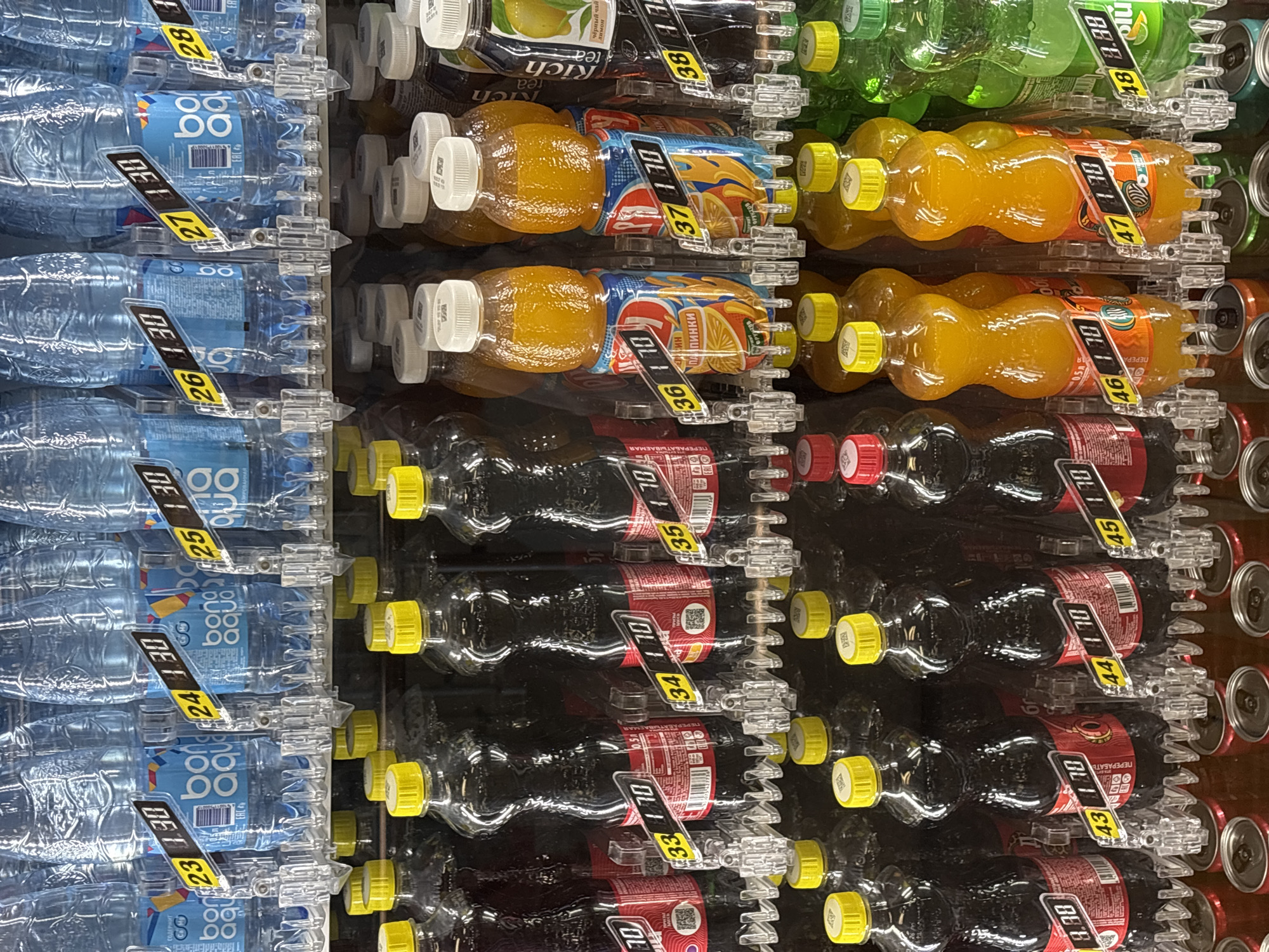 In Khostinskiy, Russia, a vending machine displays an assortment of bottled beverages. The top row features clear plastic bottles of water, some with blue labels indicating "Bona Aqua." Below, rows of drinks in various colored bottles are visible. Bottles with dark brown liquid, likely soda or cola, are present, some with red labels. Alongside these are bottles of orange-colored drinks, possibly juice or flavored beverages, with colorful labels featuring fruit imagery. Clear plastic bottles of green liquid, potentially soda or tea, are also on display in the lower section. Price tags, featuring black text on a yellow background, are attached to many of the bottles, showing prices such as "23 1.30," "24 1.30," "25 1.30," "26 1.30," "27 1.30," "28 1.30," "33 1.70," "34 1.70," "35 1.70," "36 1.70," "37 1.70," "38 1.70," "8.88," "43 1.70," "44 1.70," "45 1.70," "46 1.70," "47 1.70," and "48 8.88." The vending machine is illuminated, highlighting the products within. The background is dark, suggesting the interior of a building or a shaded area. No people are visible in the image.