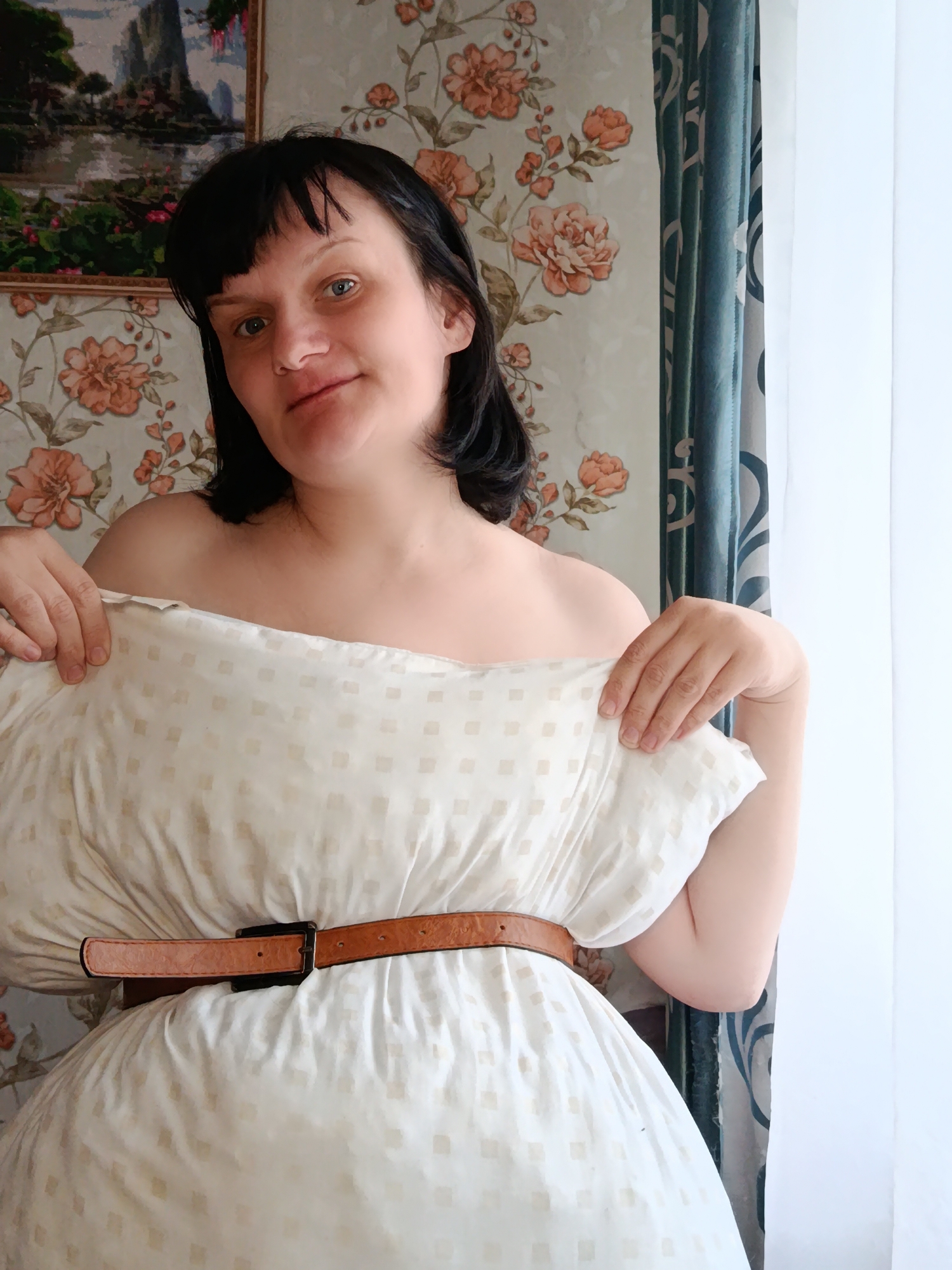A woman with dark hair and bangs looks directly at the camera with a slight smile in an indoor setting in Фоки, Russia. She is participating in a "pillow challenge," using a large, off-white pillow with a subtle square pattern as a makeshift dress. A brown leather belt with a dark buckle cinches the pillow around her waist, and she holds the top edge of the pillow with both hands, creating an off-the-shoulder effect.
Behind her, the wall is covered with light-colored wallpaper featuring a floral pattern of peachy-orange flowers. To the left, a framed picture hangs on the wall, depicting a landscape scene with mountains and water. To the right, a blue or teal curtain with a dark pattern is visible next to a bright window, suggesting it is daytime. No visible text is present in the image.