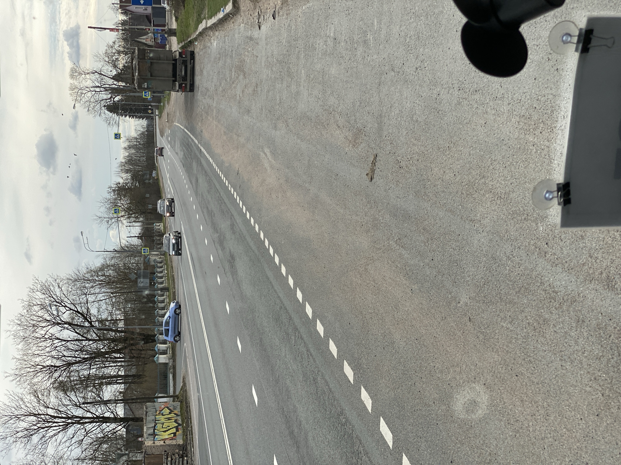 The image shows a straight, wide road with multiple lanes and dashed white lane markings. The road is gray and appears to be asphalt. Vehicles, including cars and a large truck, are visible on the road. The roadside has trees with bare branches, some buildings, and signage. Graffiti is visible on a building near the road. The photo seems to be taken from inside a vehicle, with a suction cup mount and a portion of a device visible in the foreground. The scene appears to be a road within Opolie, Russia. There are no people immediately visible.