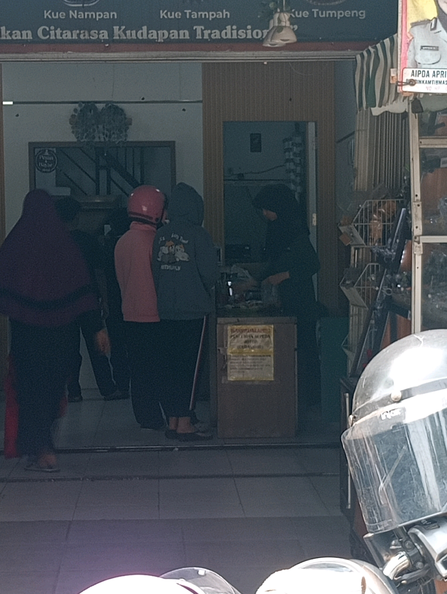 The image depicts an outdoor scene in Bandar Lampung, Indonesia, likely in front of a shop. A dark blue awning with white stripes is visible above the entrance. A sign at the top reads "Kue Nampan Kue Tampah Kue Tumpeng" and below it, "kan Citarasa Kudapan Tradisio". Further to the right, a sign is partially visible with "AIPDA APRI".
In the foreground, a person wearing a silver helmet with a clear visor is partially visible, suggesting a motorcycle is present. Inside the shop entrance, several individuals are visible. One person on the left wears a dark maroon hooded garment. To their right, another person wears a pink top and a red helmet. Next to them, a person in a dark blue hoodie with a graphic print stands. Another individual in a black hijab and long sleeves is seen behind a counter, interacting with what appears to be goods.
A display shelf with various items is visible inside the shop. A brown wooden counter or cabinet is positioned in the middle ground. On this counter, a yellow sign with text is placed, which is partially legible. The flooring appears to be tiled. The overall lighting suggests an overcast day or the shop is shaded.