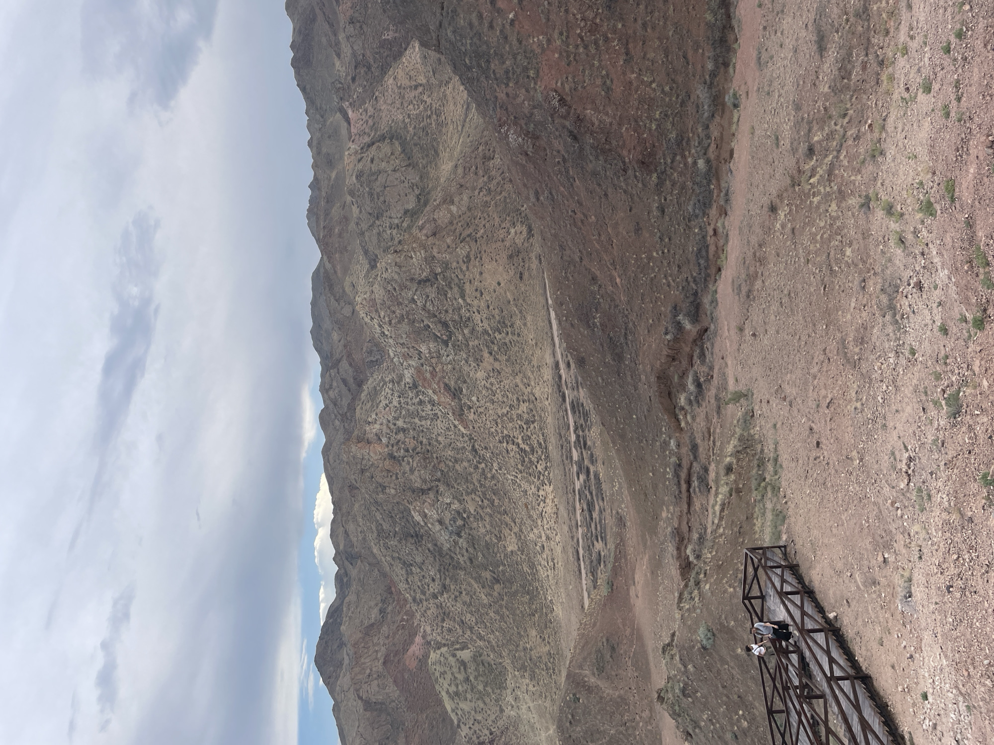 Two individuals are positioned on a wooden boardwalk that overlooks a valley. The boardwalk is made of dark wood and features a railing. The valley floor is covered in arid vegetation and scattered rocks, with a dirt road winding through it. In the background, rugged, eroded mountains rise under a cloudy sky. The landscape is predominantly earthy and muted in color, with reddish-brown hues dominating the foreground and the mountains. The individuals appear to be standing and observing the scenery. The overall impression is of a remote, natural environment with visitor infrastructure.