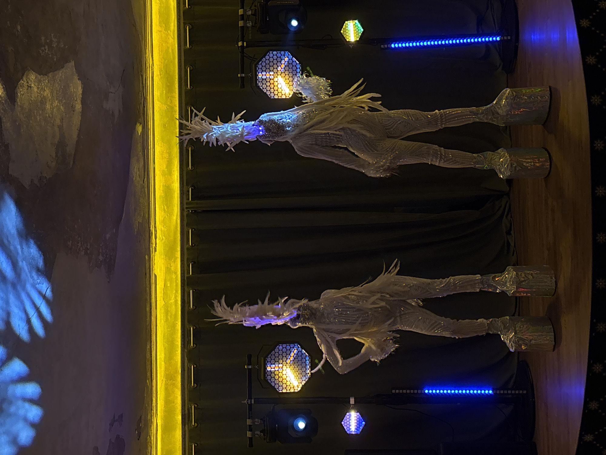 The image shows two mannequins dressed in white, elaborate costumes with feathered headdresses and platform boots. They are illuminated by various stage lights, including LED bars and hexagonal spotlights. The mannequins are positioned against a backdrop of dark curtains. A textured wall with yellow trim is visible on the left side of the image. The photo was taken indoors, likely in a performance or event space. The location is unknown.