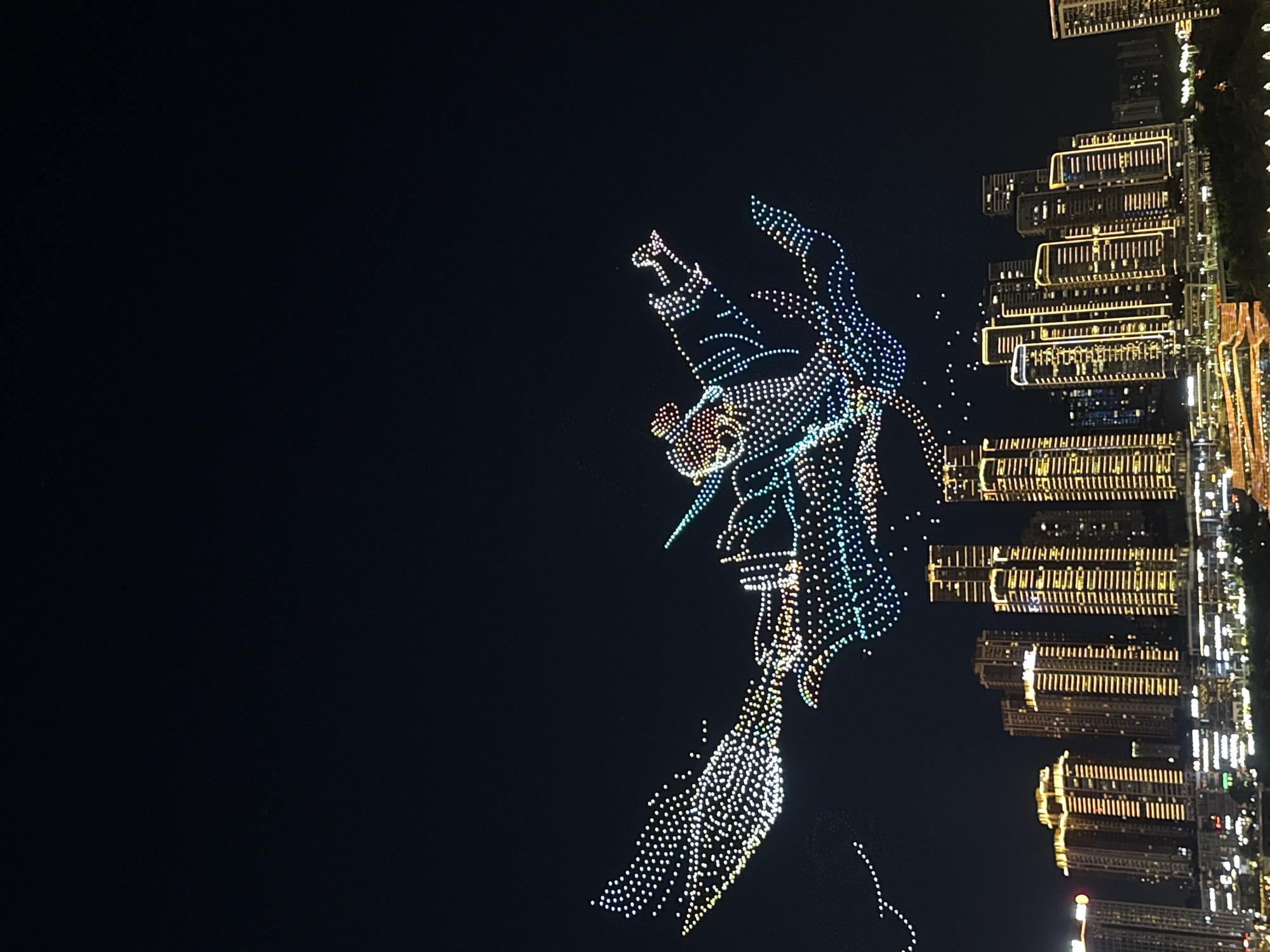 A drone show is taking place in the night sky over a cityscape, likely in Yuzhong District, China. Above the city, numerous lights form the shape of a figure, possibly a mythical creature or a character in traditional Chinese attire, with large outstretched wings. The drone formations exhibit a variety of colors, including white, yellow, green, blue, and pink. Below the aerial display, a densely packed urban landscape is illuminated. Tall buildings with golden lights outline their structures and windows dominate the skyline. Some buildings appear to be illuminated from within, while others have decorative lighting along their facades. The city extends into the distance, with more buildings visible, though less distinct due to the darkness and distance. A hint of green foliage can be seen in the foreground, suggesting a waterfront or park area. The overall scene captures a vibrant and technologically advanced nighttime event.