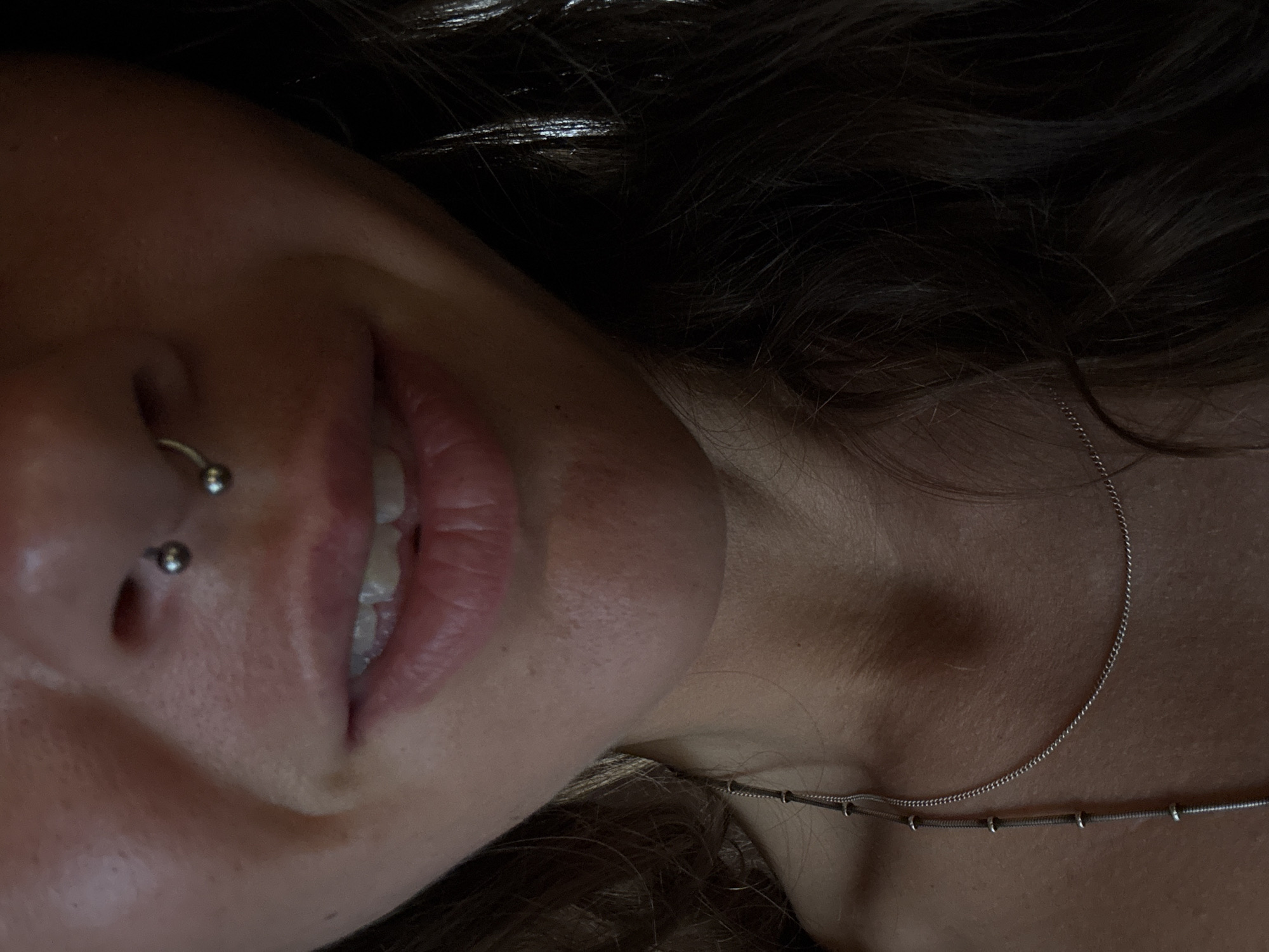 A close-up, dimly lit, eye-level shot shows a person's face from the nose down, with a portion of their chest and hair visible. The person has a septum piercing with a silver circular barbell. Their mouth is slightly open, revealing their teeth and pink lips. A delicate silver necklace with small spherical charms is visible on their collarbone. The background is dark and out of focus, with strands of dark, wavy hair on the right side of the frame.