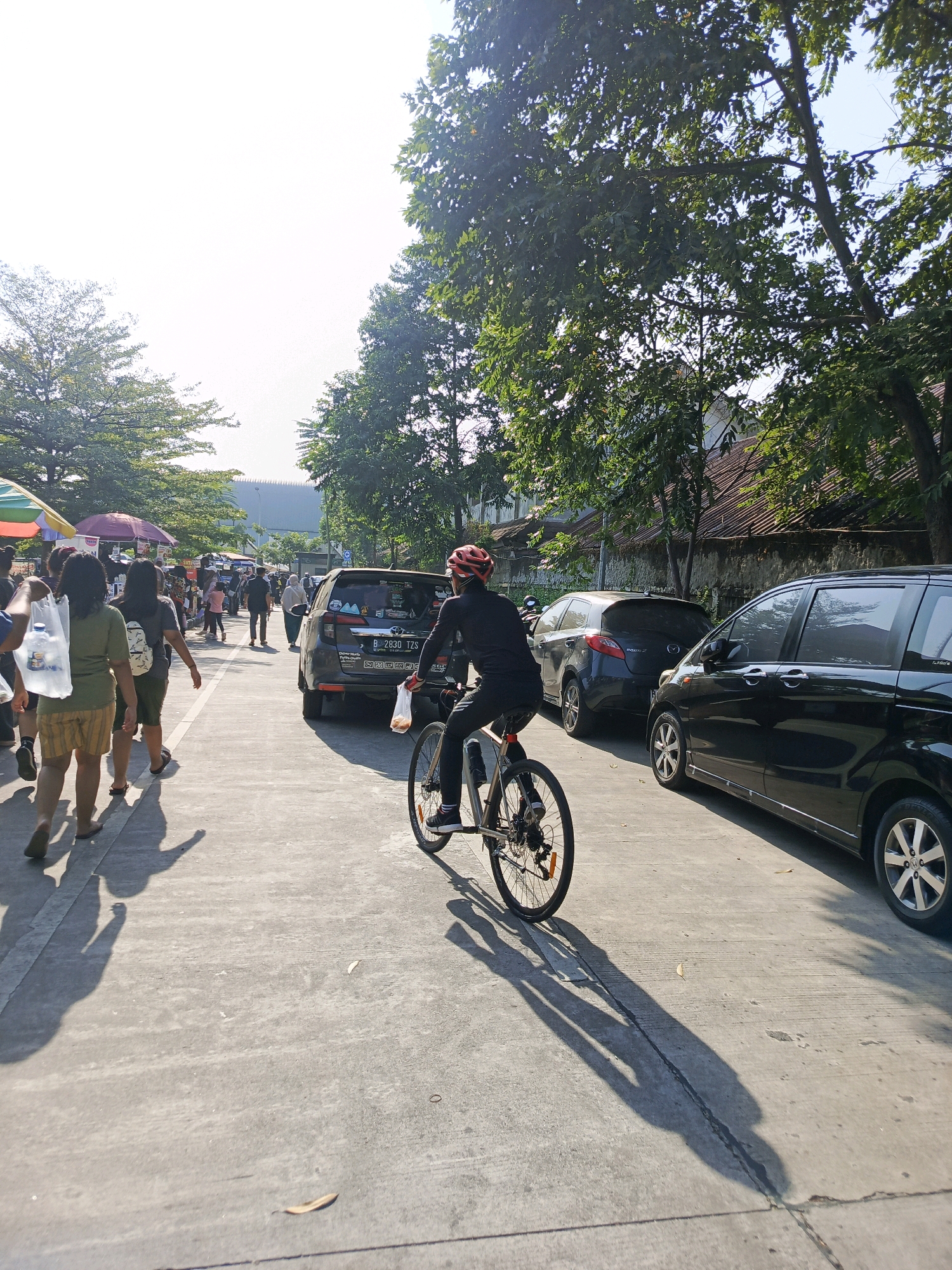 The image depicts a sunny outdoor scene in a city, likely a street market or a public thoroughfare in Tangerang, Indonesia. In the foreground, a person wearing a red helmet and black clothing is riding a bicycle. The cyclist has a plastic bag in their right hand, and the bicycle has a water bottle holder. The cyclist's shadow stretches across the concrete road.
To the left of the cyclist, several people are walking. Two women are prominent in the middle ground; one wears a green t-shirt and striped shorts, carrying a white plastic bag. The other, to her left, wears a dark t-shirt and has a backpack. Behind them, other pedestrians are visible, some carrying shopping bags. Several market stalls with colorful umbrellas are set up along the left side of the road.
Several vehicles are parked or driving on the road. A dark-colored SUV is in the mid-ground, with a license plate starting with "B 2830 TZS". To the right of the SUV, a dark hatchback is parked, and further to the right, a black minivan is parked. The background shows trees lining the street, buildings, and a bright, overcast sky. The overall impression is a busy, daytime scene with people engaged in shopping and transit.