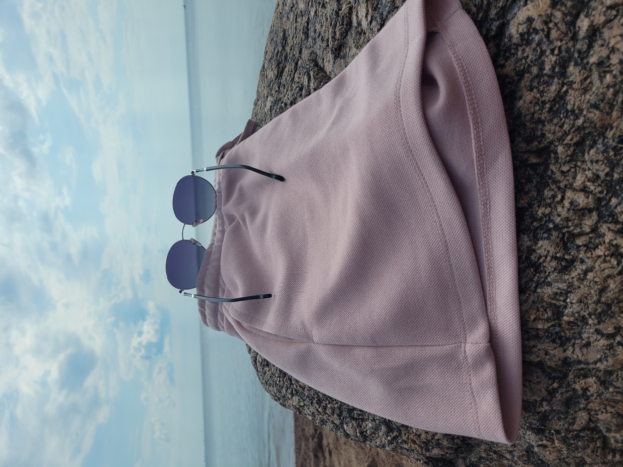 On a granite rock by the sea in Zelenogorsk, Russia, a pair of dark-rimmed, oval sunglasses rests on a pastel pink pair of shorts. The shorts are made of a textured knit fabric and are folded at the hem. The sea stretches out to the horizon under a partly cloudy sky.
