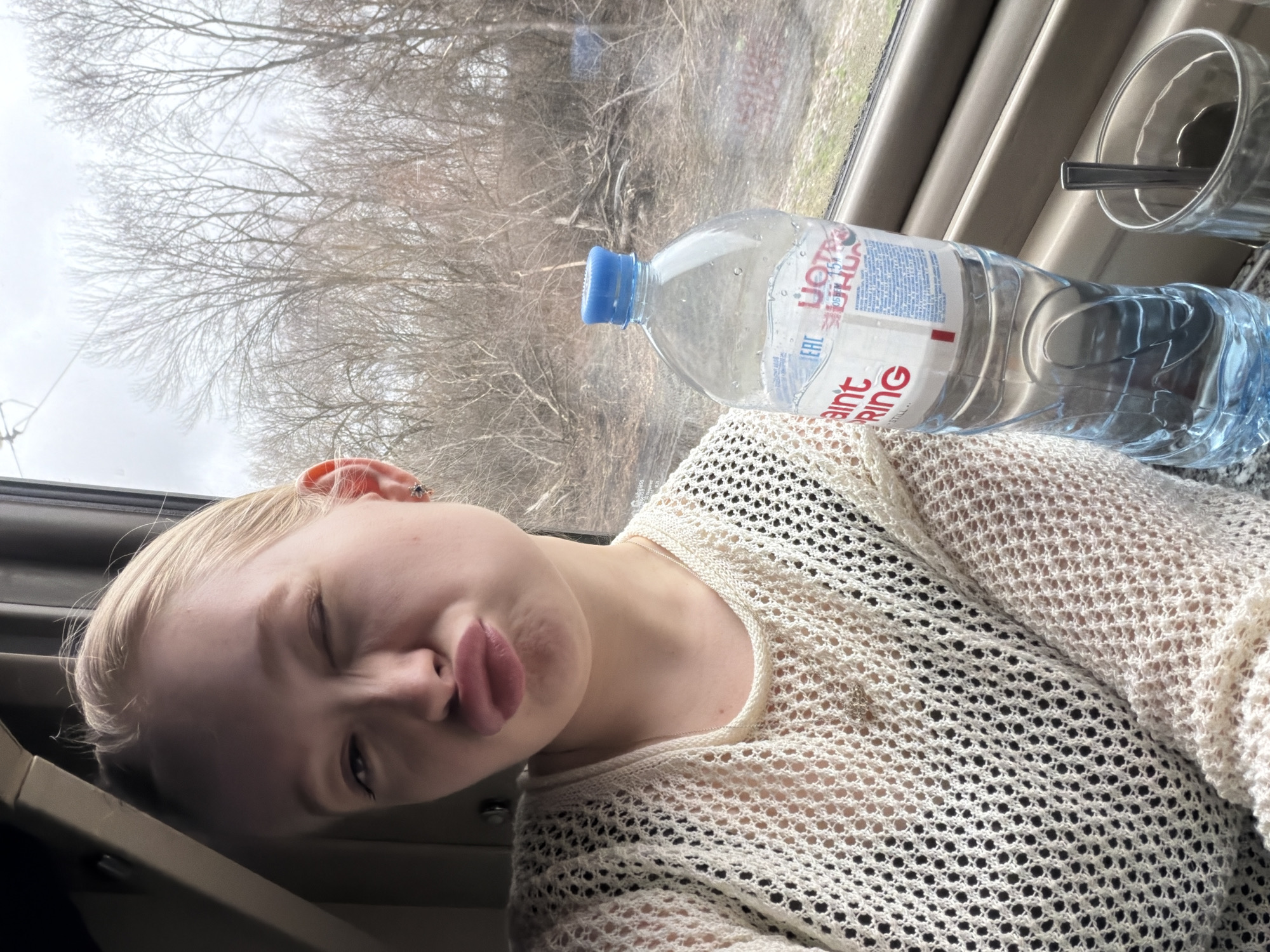 A young, fair-skinned woman with blond hair is pictured inside a vehicle, possibly a train, judging by the window and interior paneling. She is turned towards the camera with one eye closed in a playful wink and her lips pursed. She is wearing a light-colored, loosely knitted top over a black shirt. A partially filled water bottle labeled "Saint Spring" is positioned beside her on a surface near the window, along with a glass containing a spoon. Through the window, leafless trees are visible against an overcast sky. The location is likely near Зелёный Дол, Russia, based on the Cyrillic text on the water bottle and its potential availability within the region.