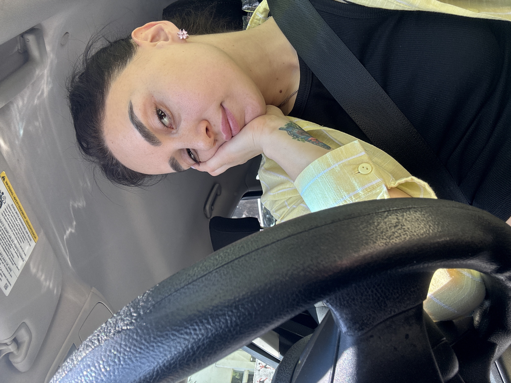 The image displays a woman seated inside a car, likely in Tbilisi, Georgia, captured during daylight hours. She is positioned towards the left of the frame, looking at the camera with a calm, slightly melancholic expression, her chin resting on her right hand.
The woman has dark hair and prominent eyebrows. She wears a small, light pink, flower-shaped stud earring in her left ear. Her clothing includes a black ribbed tank top worn under a light yellow and white plaid collared shirt. A colorful tattoo, resembling a butterfly, is visible on her right forearm/wrist. A black seatbelt is fastened across her chest.
The car's interior features a black, textured steering wheel prominent in the foreground, and a light gray headliner/ceiling. Sunlight creates bright reflections and patterned light across the ceiling and steering wheel, possibly indicating light filtered through leaves or branches outside. A sun visor on the driver's side is visible, bearing a multi-lingual warning label. Visible text from the label, confirmed by OCR, includes "EVEN WITH ADVANCED AIR BAGS," "WARNING/AVERTISSEMENT/ADVERTENCIA," "Always use seat belts and," "See Owner's Manual," "ACS GONFLABLES PERFECTIONNÉS" (French for "Advanced Airbags"), and "BOLSAS DE AIRE" (Spanish for "Airbags"). Through the steering wheel, glimpses of the exterior, including what appears to be part of another vehicle or building, are visible. The overall setting suggests a casual moment while in transit or parked.