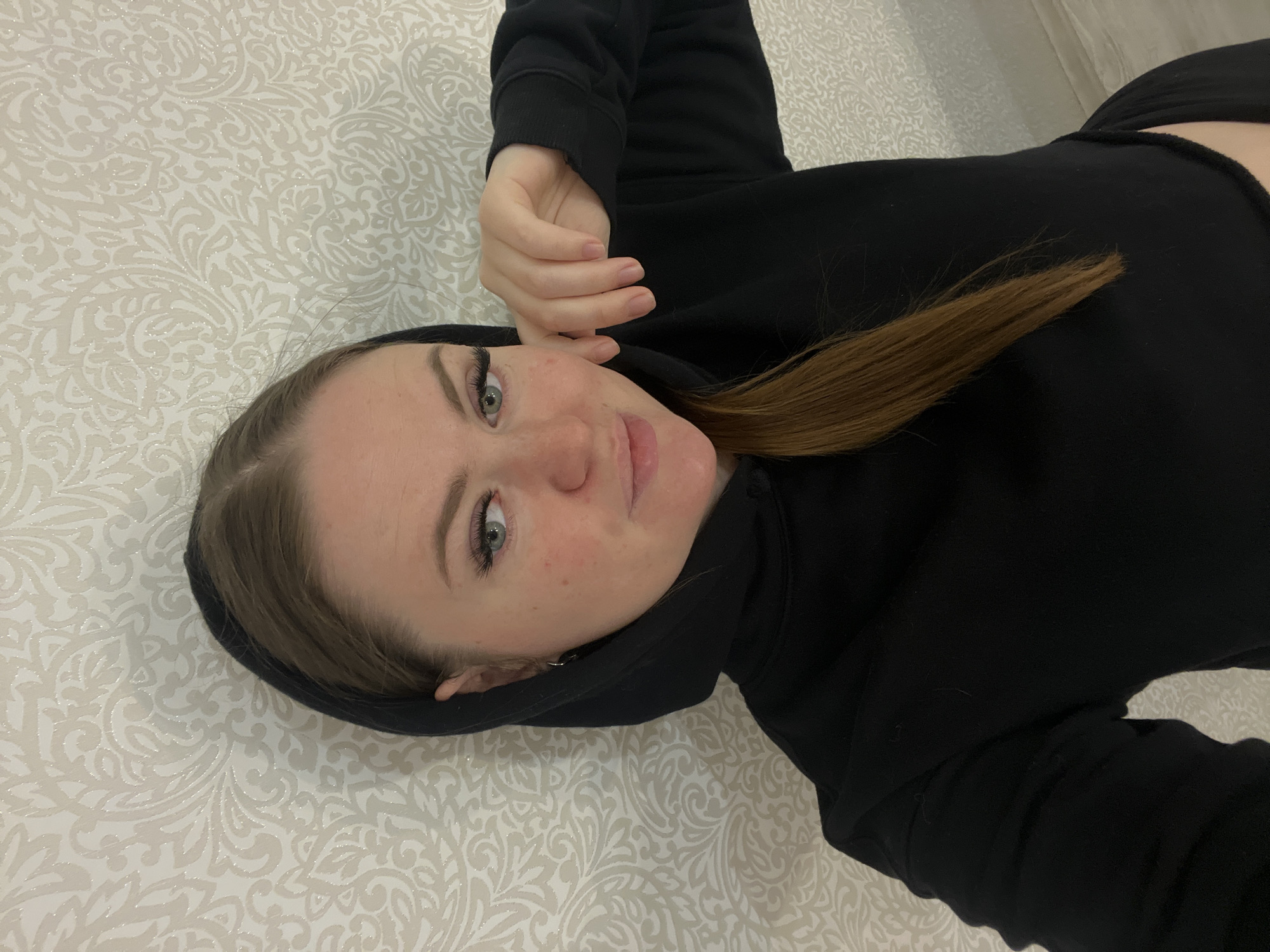 A woman lies on a patterned surface, likely a bed. She wears a black hooded sweatshirt. The woman's hair is light brown. The background is a neutral-toned patterned wallpaper or bedspread. The location is not discernible from the image. There is no indication of the city of Sevastopol, Ukraine.