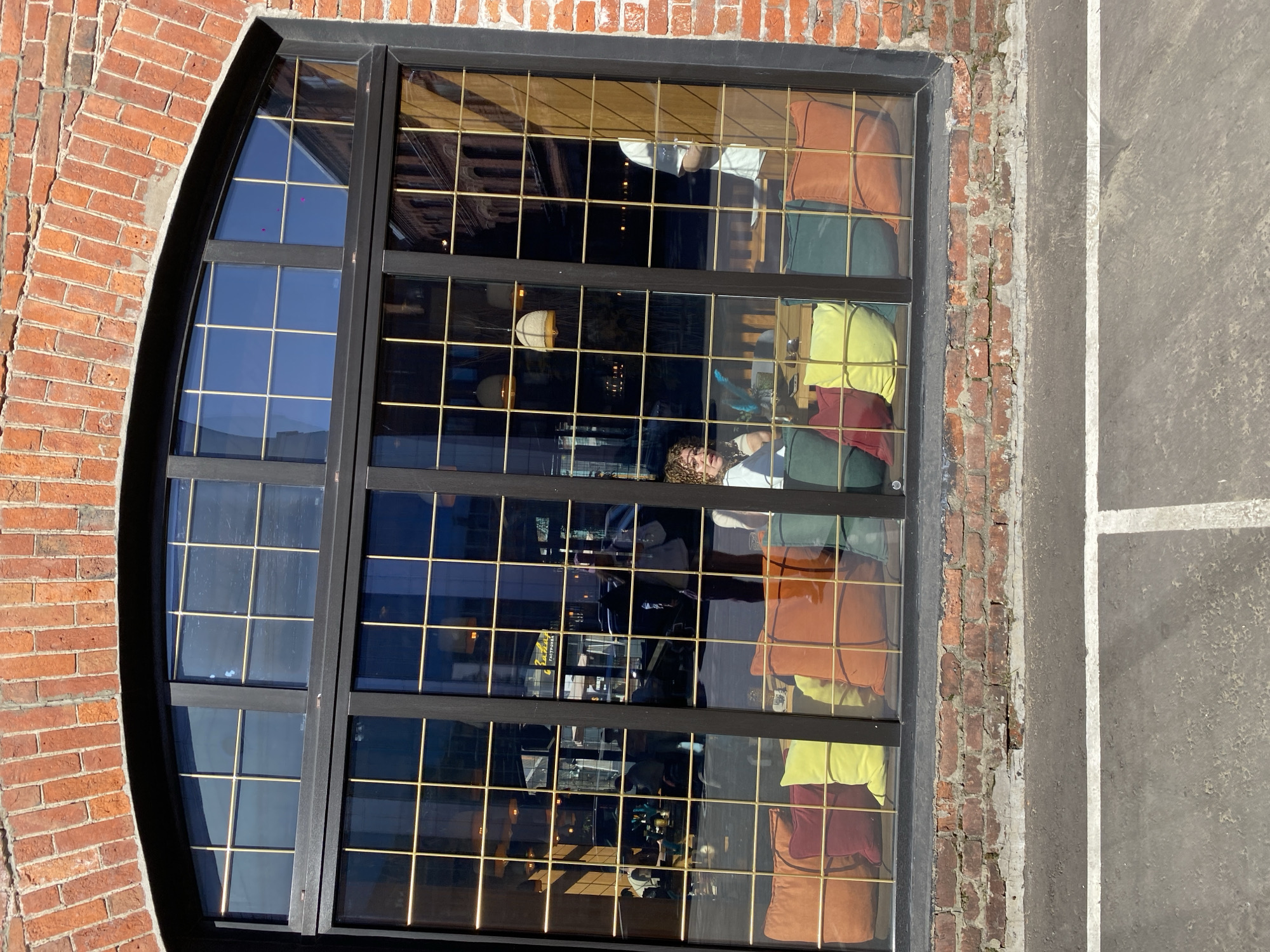 Here is a description of the image:
The image shows a large, multi-paned window set into a brick building. The window has a black frame and the left portion is arched at the top, mirroring the brickwork above. The window is reflecting the interior of what appears to be a restaurant or cafe. Inside, there are colorful pillows, furniture, and lighting fixtures. A person with curly hair is visible near the center of the reflection, along with other blurred figures. A parking area or street is visible on the right side of the image. The architectural style and brickwork suggest an urban setting. The restaurant is located in Moscow, Russia based on visual information.
