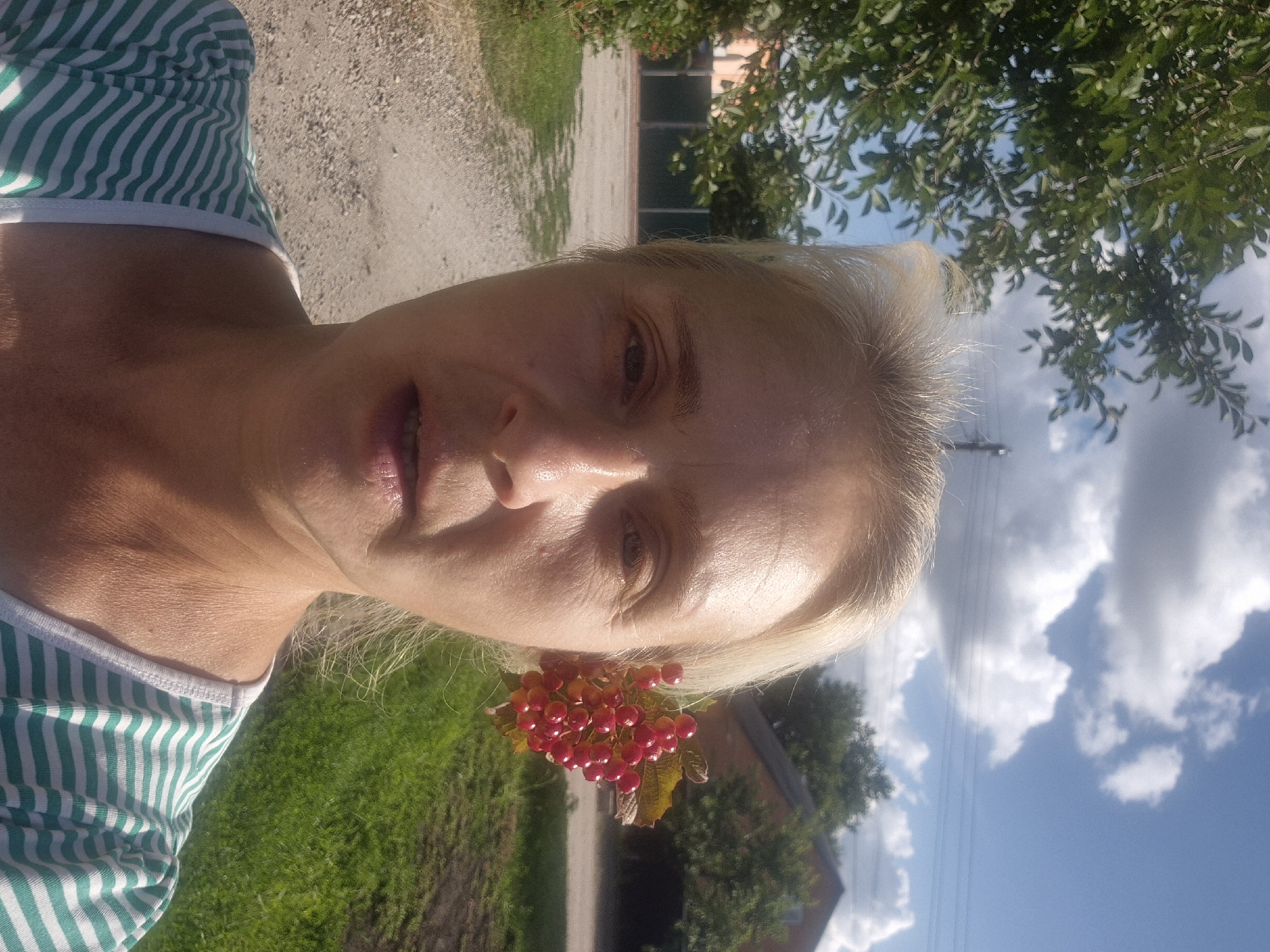 A medium shot shows a fair-skinned woman with blond hair, wearing a green and white striped t-shirt. A sprig of red berries is tucked behind her right ear. She is standing outdoors, with a gravel path and green grass visible in the foreground, and trees and a house in the background. The sky is blue with white clouds. The setting is likely the village of Sozonivka, Ukraine, given the rural environment.