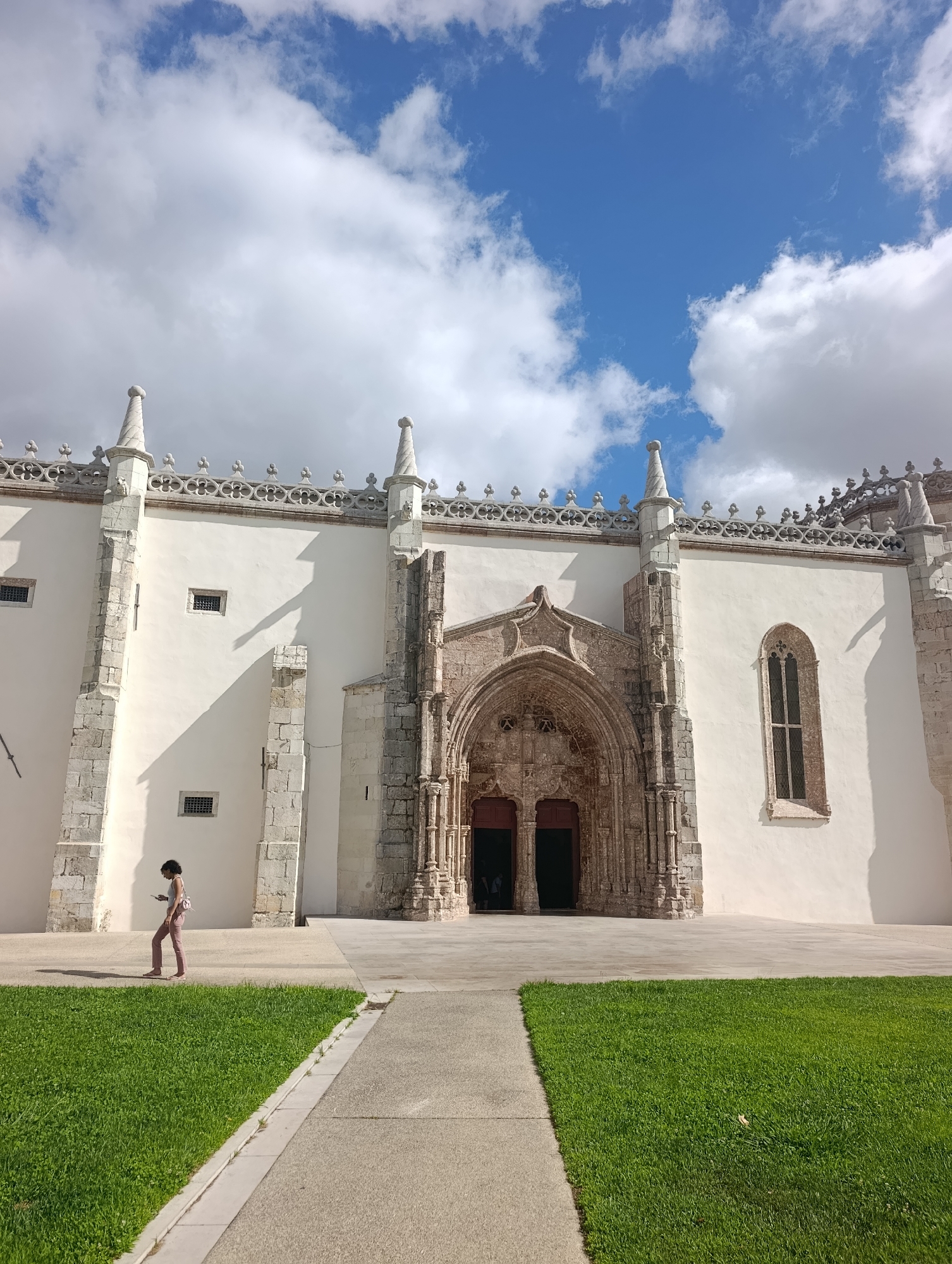The image shows the Jerónimos Monastery in Lisbon, Portugal, not Setúbal. A person is walking on a pathway in front of the monastery's ornate entrance. The person is wearing pink trousers and a light-colored top, carrying a small bag. The monastery facade is white with intricate stonework and decorative pinnacles along the roofline. The sky above is blue with scattered white clouds. Green grass and paved walkways form the foreground and surrounding area.