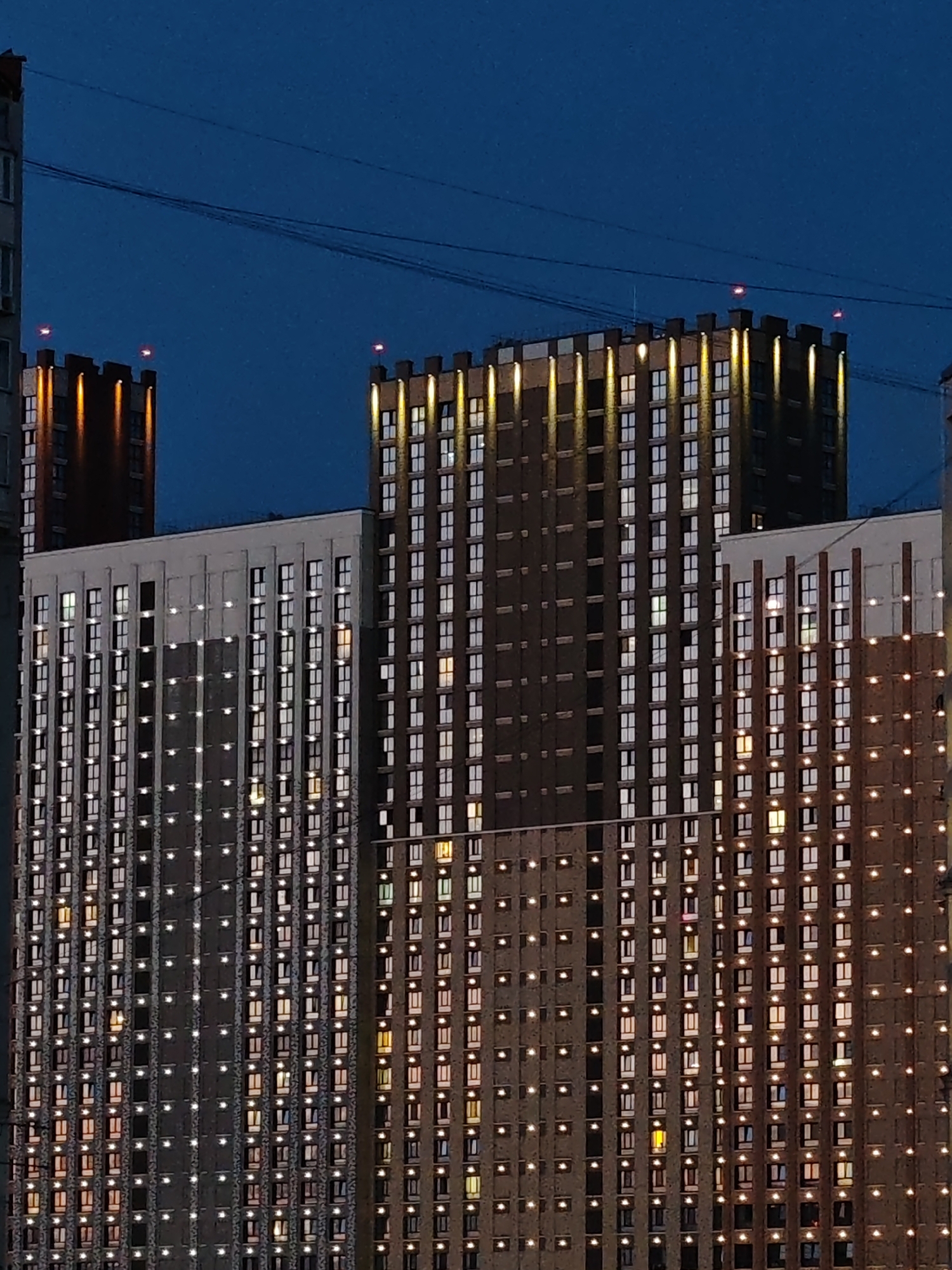 A nighttime, exterior, vertical, medium shot captures the illuminated windows of several high-rise apartment buildings against a deep blue sky. The buildings are densely packed with rectangular windows, many of which are lit from within, creating a grid-like pattern of light. The buildings have a mix of dark and lighter colored facades, with some featuring decorative lighting strips along their upper edges. Power lines are visible stretched across the sky. The scene is devoid of any people or discernible activity. The location is listed as Unknown, Unknown.