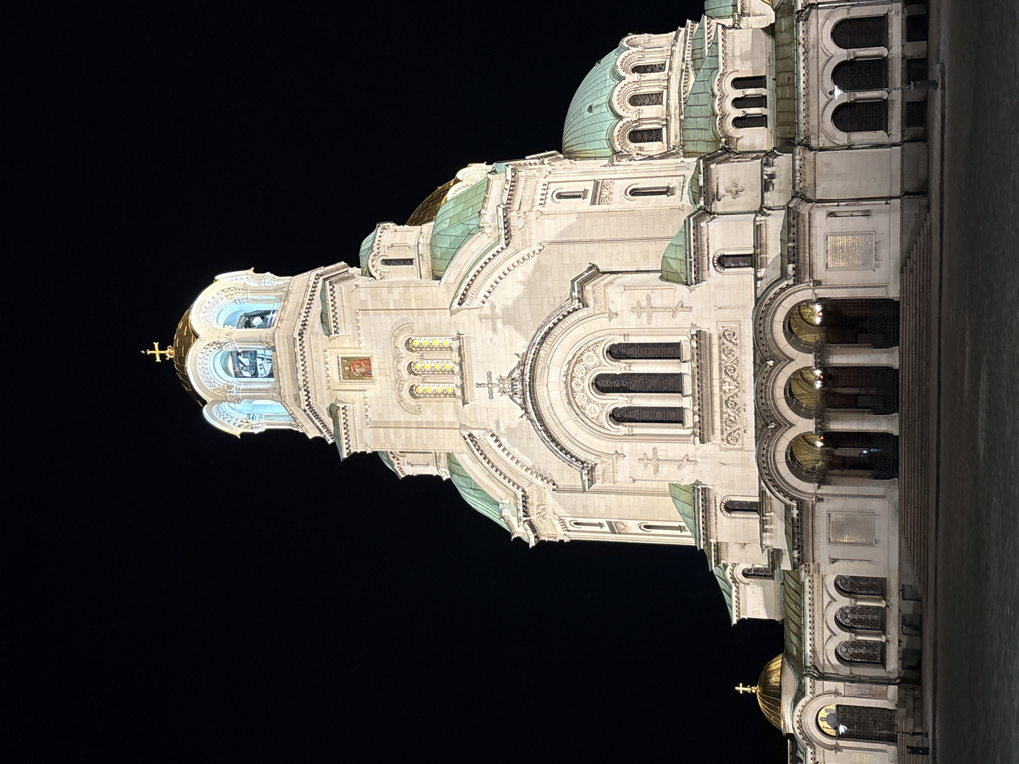 The Alexander Nevsky Cathedral is seen at night in Sofia, Bulgaria. The cathedral is ornately designed with several domes and arched windows. The architecture is illuminated with white light, creating a stark contrast against the dark sky.