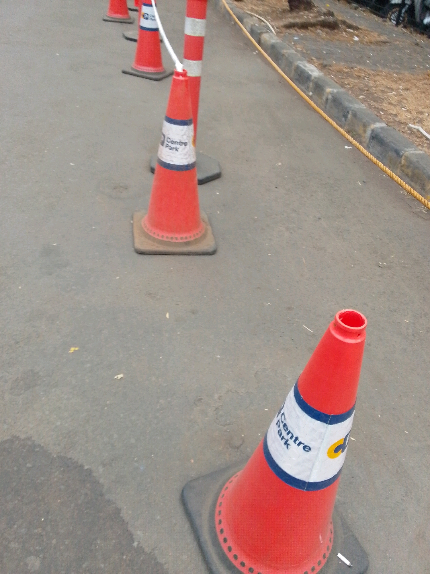 A line of orange traffic cones marks a path on an asphalt surface. The cones are spaced at intervals, and some have white reflective bands with blue lettering that reads "Centre Park". A yellow rope runs along a concrete curb to the right of the cones, separating the asphalt from a dirt and debris-filled area. In the background, a motorcycle is partially visible. The scene appears to be outdoors, possibly in a parking lot or along a street in East Jakarta, Indonesia.