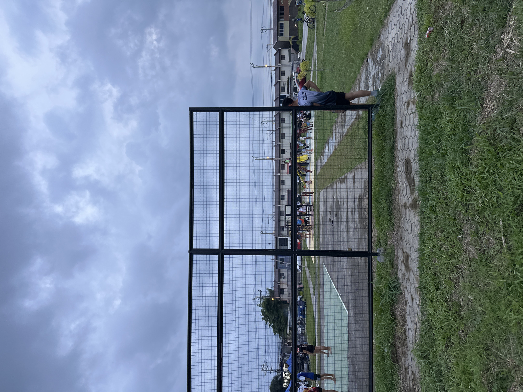 The image depicts an outdoor recreational area, likely a sports court, in Davao City, Philippines, under a heavily overcast sky. A tall black metal wire fence dominates the foreground, dividing the scene.
On the right side of the fence, a person wearing a grey short-sleeved shirt and black shorts is leaning against the fence, facing away from the camera. The back of their shirt clearly shows two characters: "古口". Adjacent to the fence on the right is a paved path made of hexagonal concrete blocks, bordered by lush green grass. A discarded red can is visible lying on the grass near the path.
Behind the fence, a green-painted sports court or field is visible, where several individuals are gathered, some appearing to be engaged in activities or socializing. In the middle ground, residential buildings with multiple stories are visible, along with utility poles and power lines crisscrossing the sky. A bicycle is parked near one of the buildings. The sky is filled with thick, grey clouds, suggesting either an impending rain or an overcast late afternoon. The overall impression is one of a typical community recreational space.