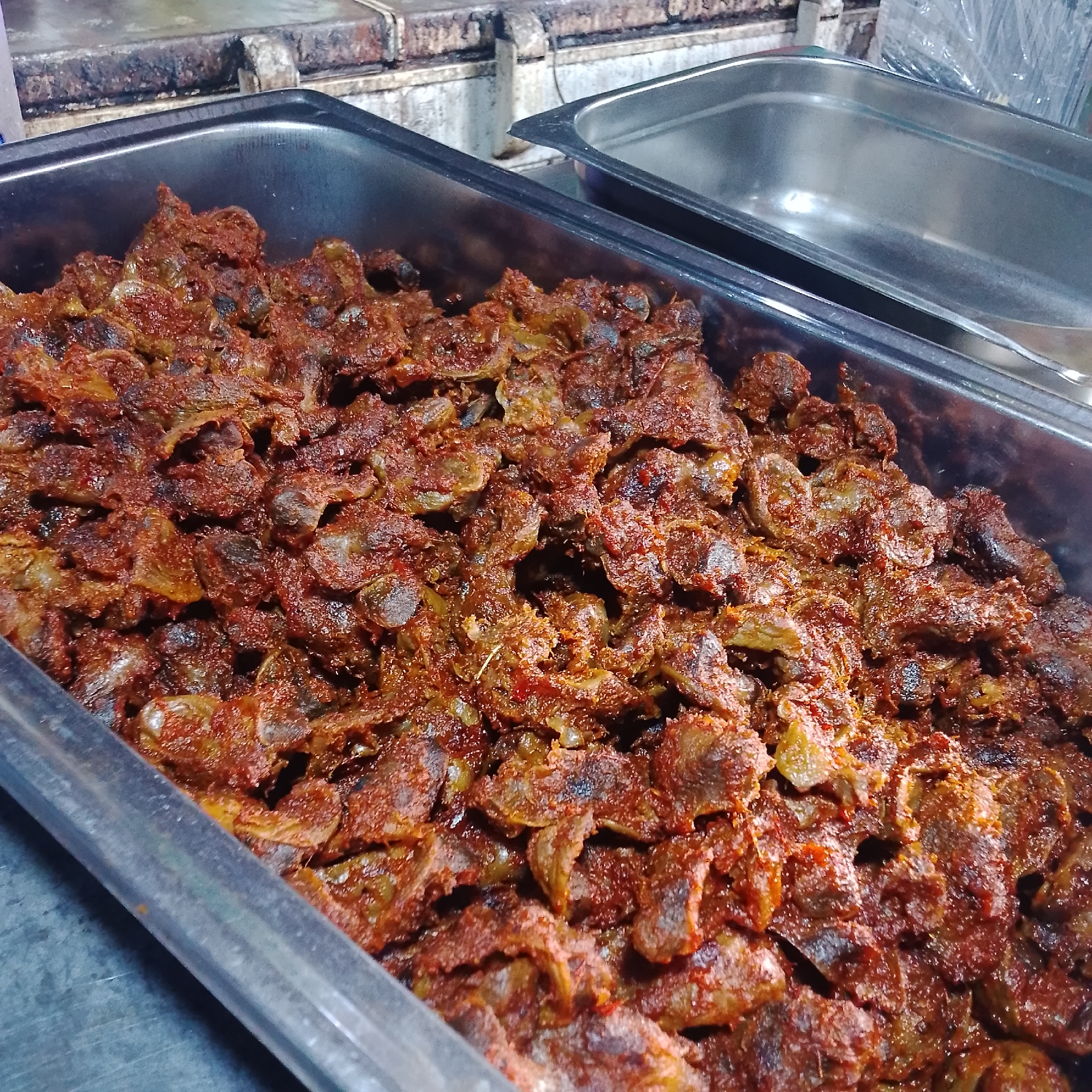 A close-up shot shows a large, rectangular metal serving tray filled with a generous portion of small, dark brown, and reddish pieces of meat, likely chicken gizzards, coated in a thick, spicy seasoning. The meat appears to be stir-fried or braised, with some pieces exhibiting a slightly charred texture. The seasoning is a rich, earthy red, suggesting ingredients like chili peppers, tomatoes, and various spices. A second, empty metal serving tray is visible to the right of the main tray, placed slightly behind and to the side. The trays are situated on a counter or table surface, which appears to be made of a dull metal. In the background, the image captures elements of a food stall or restaurant setting, with what looks like a stainless steel kitchen range or preparation area behind the serving trays. The overall impression is of a food preparation area in Lagos, Nigeria, where this dish is ready to be served.