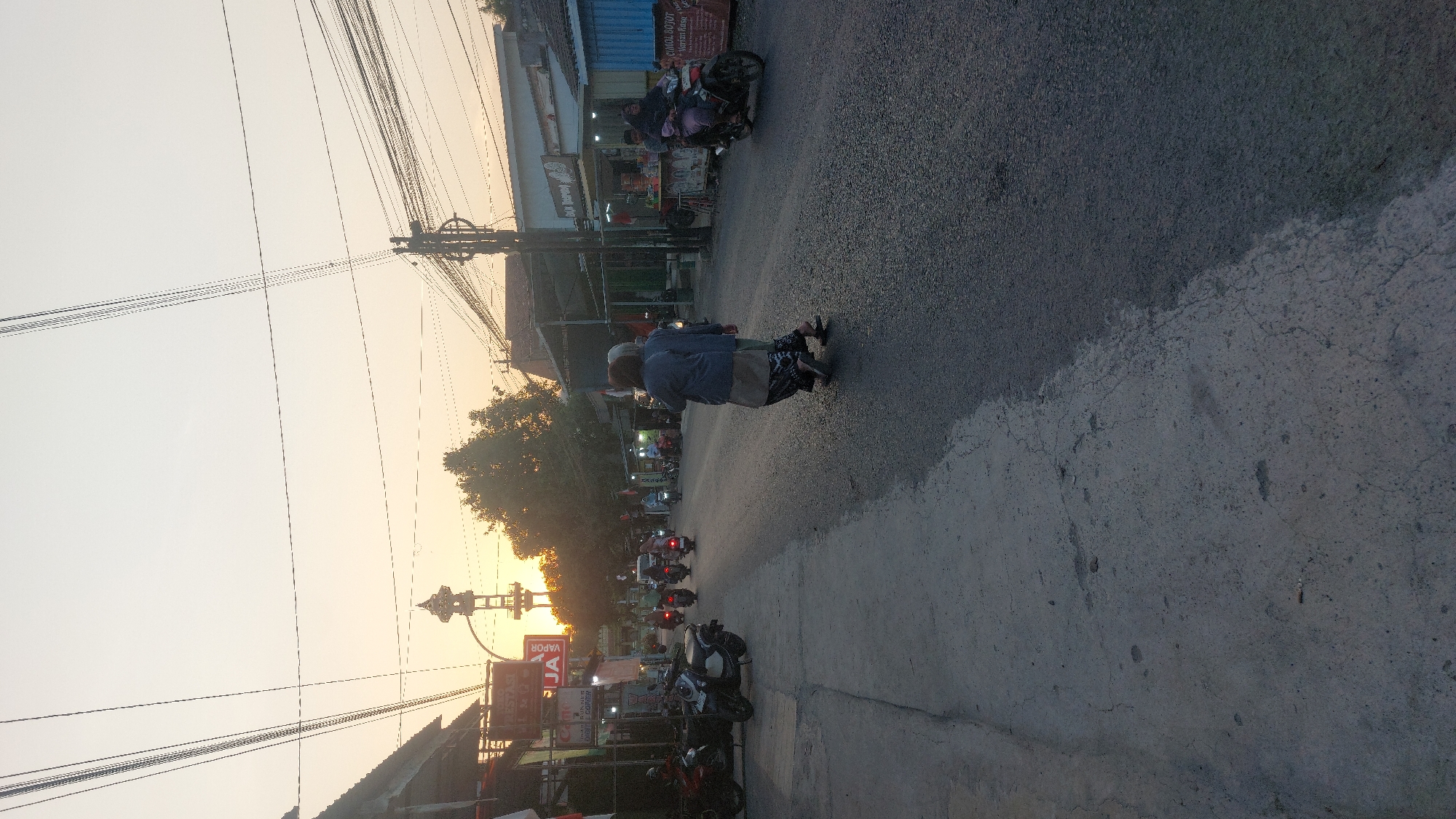The image depicts a street scene in the city of Karas, Indonesia, during what appears to be sunset or dawn. The sky is a soft orange and yellow, with power lines crisscrossing overhead. On the left side of the street, there are several motorcycles parked on a concrete area, and buildings with signs are visible. The road itself is paved and appears to be a two-lane street with traffic.
In the mid-ground, two people, likely women based on their attire, are walking across the street from right to left. They are wearing traditional Indonesian clothing, including headscarves and long skirts. Further down the street, several motorcycles are in motion, with riders. The buildings on the right side of the street are also visible, with some storefronts and awnings. One building has a sign that reads "DUM BEBEQ" and another sign with Indonesian text. There is also a motorcycle carrying two people, a rider and a passenger, heading towards the viewer. The overall atmosphere is that of a typical Indonesian street during the evening hours, with a mix of pedestrian and vehicular traffic. The presence of the tower-like structure in the distance, a common sight in Indonesian towns, further anchors the location.
