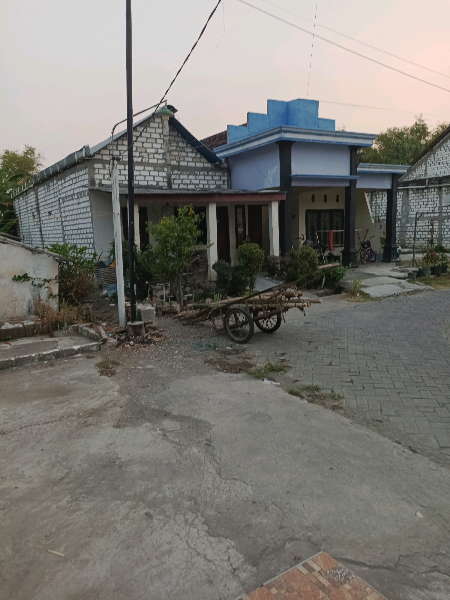 The image depicts a street scene in Rancang Kencono, Indonesia. In the foreground, a concrete pathway transitions to a paved area constructed with interlocking bricks. A traditional wooden cart, laden with bamboo poles, is positioned on the paved area. Adjacent to the paved area is a concrete pathway, part of which is visible in the lower right corner with decorative tilework.
The middle ground features two houses. The house on the left is partially constructed with exposed white bricks and a corrugated metal roof. A dark pole, likely a utility pole, stands in front of this house, with electrical wires extending across the scene. In front of the house on the right, a covered porch area with dark pillars is visible, leading into the house. A bicycle is leaning against one of the pillars. The area between the houses and the road appears unkempt, with scattered debris and vegetation.
In the background, more houses with exposed brickwork are visible. Trees and foliage are present on the right side of the scene. The sky is a pale, overcast hue, suggesting either early morning or late afternoon. The overall impression is of a residential area undergoing some level of development or in a state of disrepair.