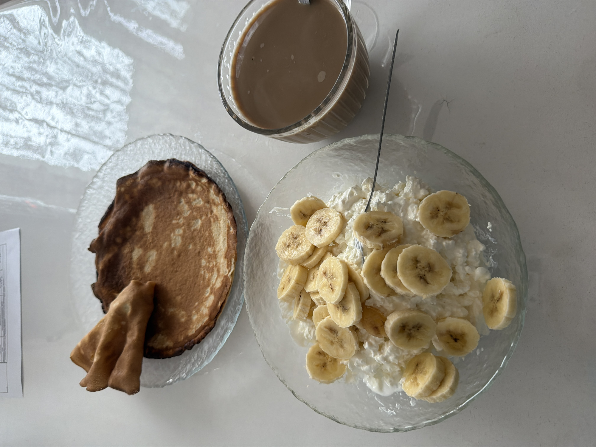 The image presents an overhead view of a meal laid out on a white countertop. A clear glass mug filled with a light brown liquid, likely coffee with milk, is positioned at the top. To the left, a clear glass plate holds several pancakes, some folded. To the right, a clear glass bowl contains cottage cheese topped with sliced bananas, and a fork is placed in the bowl. A sheet of paper is visible at the left edge of the scene. There is no indication of the city of Leningradskaya, Russia, or of any people.