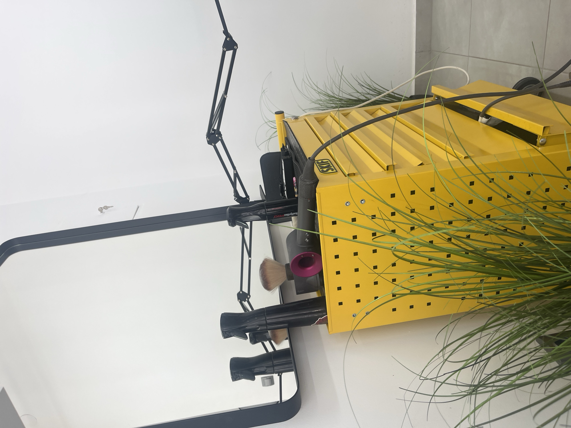 The image shows a bright yellow, multi-drawer tool cart with various hair styling tools on top. These include two black spray bottles, a makeup brush, a Dyson hairdryer, and a curling iron. A black metal articulating lamp extends over the cart. In the background, a large, rectangular mirror with a dark frame is leaned against a white wall. To the left of the cart, a few fronds of artificial green grass are visible. A small key is hanging on the white wall to the left of the mirror. The floor appears to be tiled in a light gray color. The scene is indoors, likely in a salon or barbershop. There are no people present. The image does not provide any information about the city of Uzhhorod, Ukraine, or any details that would place it in that specific location.