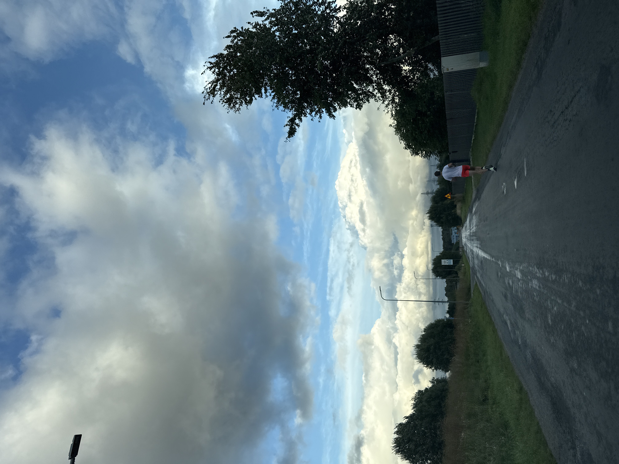 The image shows a person running on a wet asphalt road in Järveküla, Estonia. The sky is cloudy with patches of blue. There are trees and grass on the sides of the road, and a wooden fence on the right. A street lamp is visible in the upper left corner. The road appears to be in a rural or suburban area. The person is wearing a white t-shirt and red shorts. The scene suggests it may have recently rained.