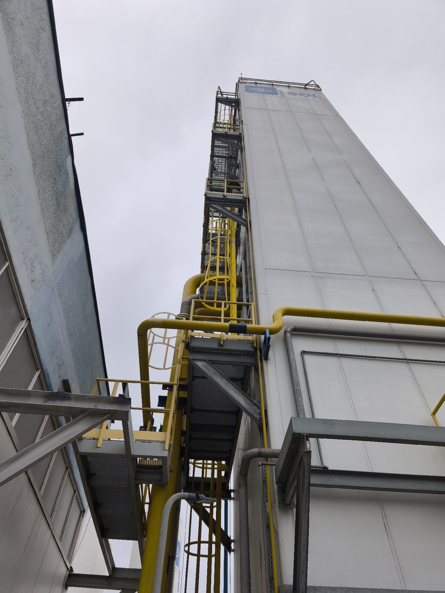 An upward-facing, low-angle shot captures a towering industrial structure alongside a shorter building under an overcast sky. The dominant structure on the right is a tall, white or light grey, panelled building, possibly a silo or storage tank, with the blue word "SOL" and a logo (resembling an eye or stylized letter) prominently displayed near its top. The word "GROUP" might also be visible below "SOL".
Running vertically and horizontally along the tall structure are numerous bright yellow pipes and an intricate system of metal staircases, platforms, and caged ladders, all painted yellow or grey. These access points suggest maintenance or operational use. To the left, a smaller building with a textured, light blue and grey wall stands, with its roofline and upper section partially visible. Grey metal beams and structural elements connect or are adjacent to both buildings.
The scene appears to be an industrial or commercial setting, such as a factory, plant, or storage facility. The lighting is soft and even, indicating an overcast day, and no specific time of day beyond daylight can be determined. There is no human activity taking place, and the image conveys a sense of scale and industrial functionality.