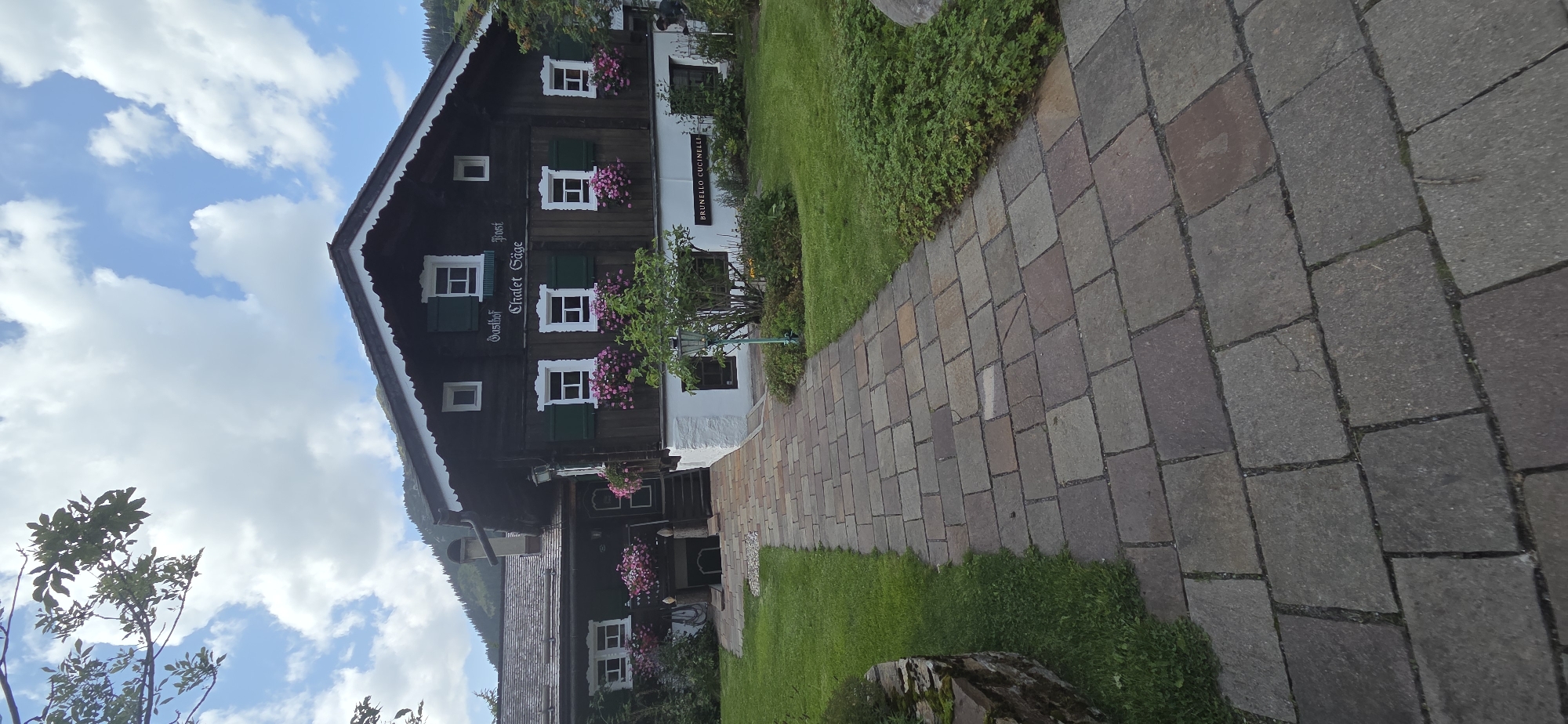 A traditional Austrian wooden chalet, identified by signage as "Gasthof Chalet Säge," is set against a backdrop of lush green hills and a partly cloudy sky. The chalet features dark wooden siding, white trim around the windows, and dark green shutters. Several windows are adorned with planters overflowing with pink flowers. A paved walkway made of irregular stone slabs leads towards the entrance, which is flanked by manicured green lawns and shrubbery. A small tree is positioned in front of the chalet. A sign on the lower part of the building reads "BRUNELLO CUCINELLI." The overall scene depicts a serene, picturesque setting typical of the Austrian Alps.