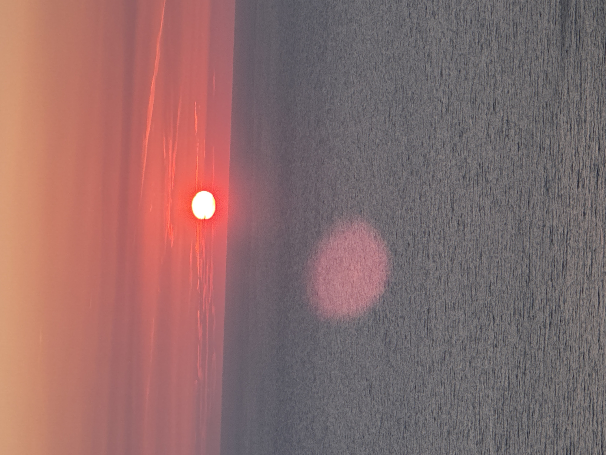 A wide-angle shot captures a sunset over the Adriatic Sea near Donji Katun, Montenegro. The sun, a bright white orb with a red halo, is positioned in the upper center of the frame, casting a pinkish glow across the sky. Wispy clouds streak horizontally, tinted with the warm hues of the setting sun. Below the sky, the sea stretches to the horizon, its surface rippled by gentle waves and reflecting the diffused light of the sun as a large, soft pink oval. The overall impression is one of serene natural beauty.