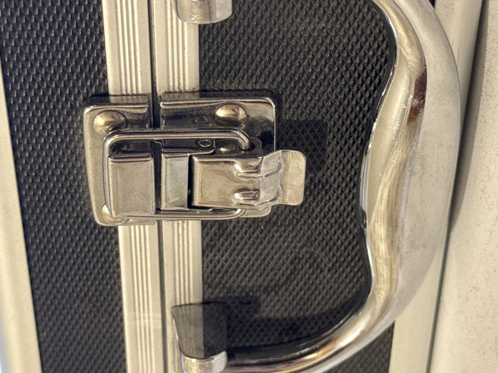 The image shows a close-up of a black case with a silver handle and latch. The case has a textured surface. The latch is made of shiny metal and is in the closed position. The handle is also made of shiny metal and is curved. The background is a light-colored surface. This image is not located in the city of Яромінскі сельскі Савет, Belarus.