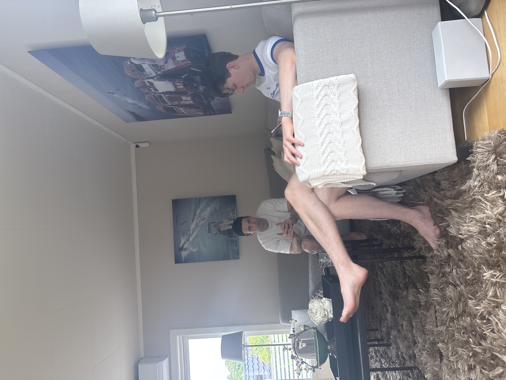 Two men are in a living room. One man is sitting on a gray sofa with a white blanket draped over his lap. He is wearing a white t-shirt with blue trim. The other man is sitting on a gray sofa behind him, wearing a white t-shirt and dark shorts. He is looking towards the camera. There is a coffee table in front of the sofa with a green bowl, a statue, and glasses on it. A floor lamp is to the right of the sofa, and a large painting of boats is on the wall behind the men. A window with a view of trees and houses is visible to the left. An air conditioning unit is mounted high on the wall near the window. The room has a light beige wall and a thick, beige shag carpet. The scene is in Larkollen, Norway.