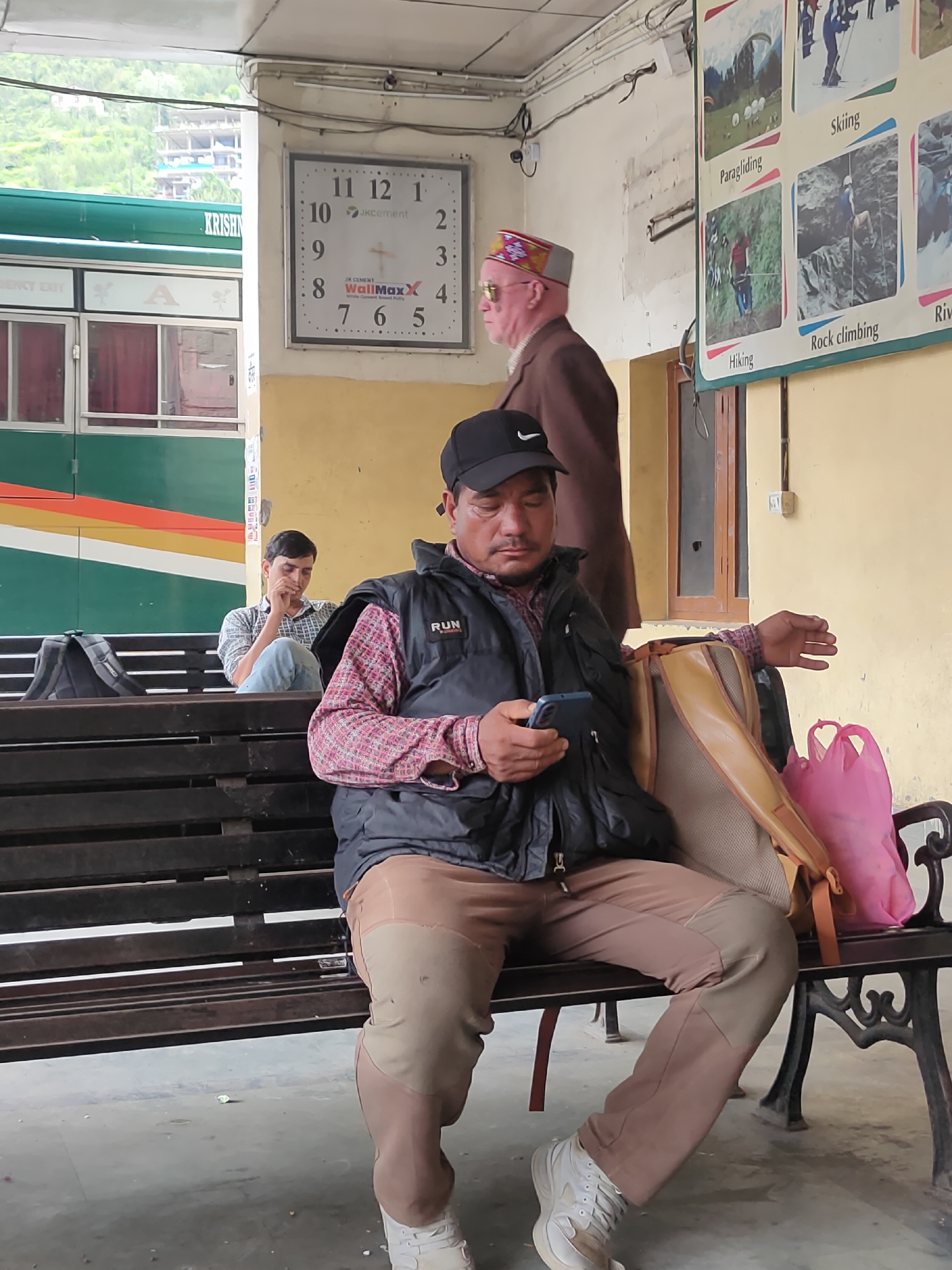 A man in a checkered shirt and a black vest sits on a dark wooden bench at a bus station in Manali, India, looking at his phone. He wears a black baseball cap and tan pants. To his left, another man in a blue and white checkered shirt and jeans sits on the same bench, looking down. Behind them, a green and white bus with orange and yellow stripes is parked. A large analog clock hangs on the wall to the left of the bus. A poster depicting various adventure sports like paragliding, skiing, hiking, and rock climbing is on the wall to the right. There are two pink plastic bags and a tan backpack on the bench next to the man on his phone. The environment appears to be an open-air waiting area with a concrete floor and a yellow-painted wall.
