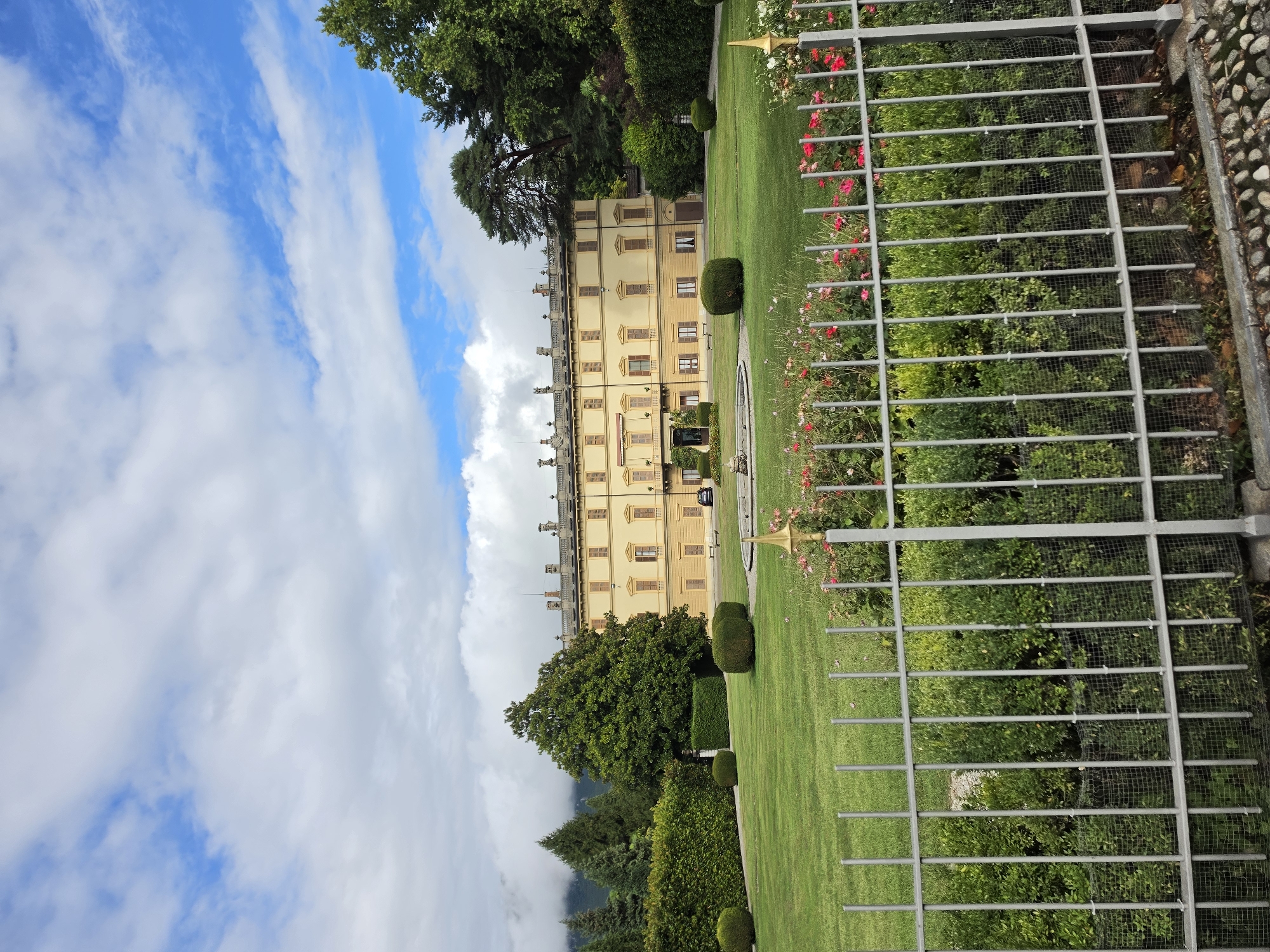 The image shows a grand yellow villa with multiple windows and brown shutters, set against a backdrop of lush green trees and a partly cloudy blue sky. The villa has a long, symmetrical facade with decorative elements along the roofline. In front of the villa, there is a well-maintained lawn with a fountain in the center. A white metal fence runs across the foreground, separating the viewer from the garden, which features manicured hedges, trimmed bushes, and a vibrant flower bed with red and pink roses. Beyond the villa, more trees and mist-covered mountains are visible. A black car is parked on the paved area in front of the villa. The scene is tranquil and appears to be an estate or a historical building, likely a tourist attraction or a private residence. The specific location in San Giovanni, Italy is not explicitly visible in the image but the architectural style and landscaping suggest a European setting.