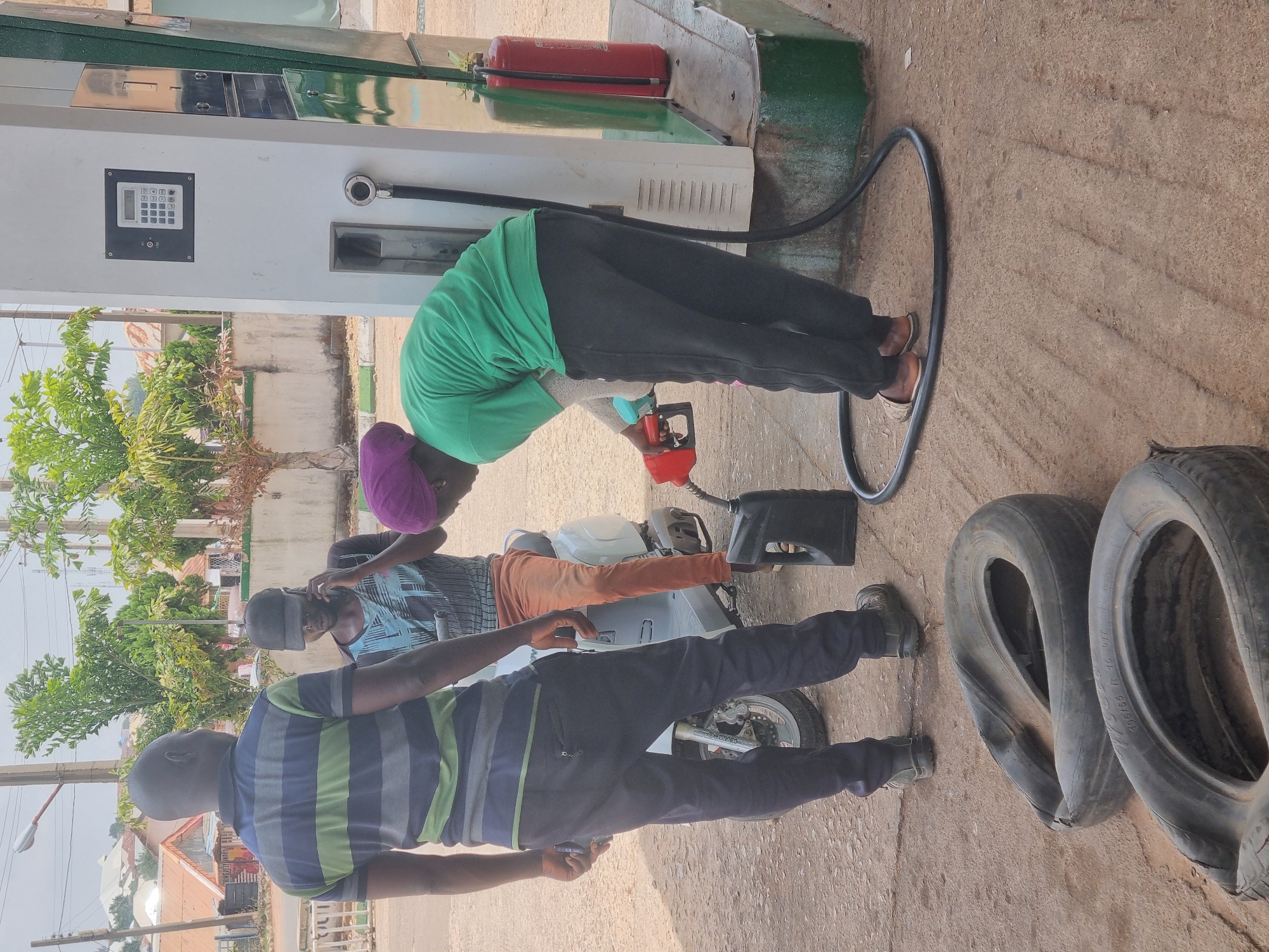 Three individuals are situated at a fuel station. One person, wearing a green shirt and a purple head covering, is filling a motorcycle's fuel tank using a red gas pump nozzle. A second individual, wearing a patterned blue shirt, appears to be seated on the motorcycle. A third person, wearing a striped blue and green shirt and black trousers, stands adjacent to the motorcycle. Two tires are placed near the lower right corner of the image. The fuel station is located in the city of Jos, Nigeria.