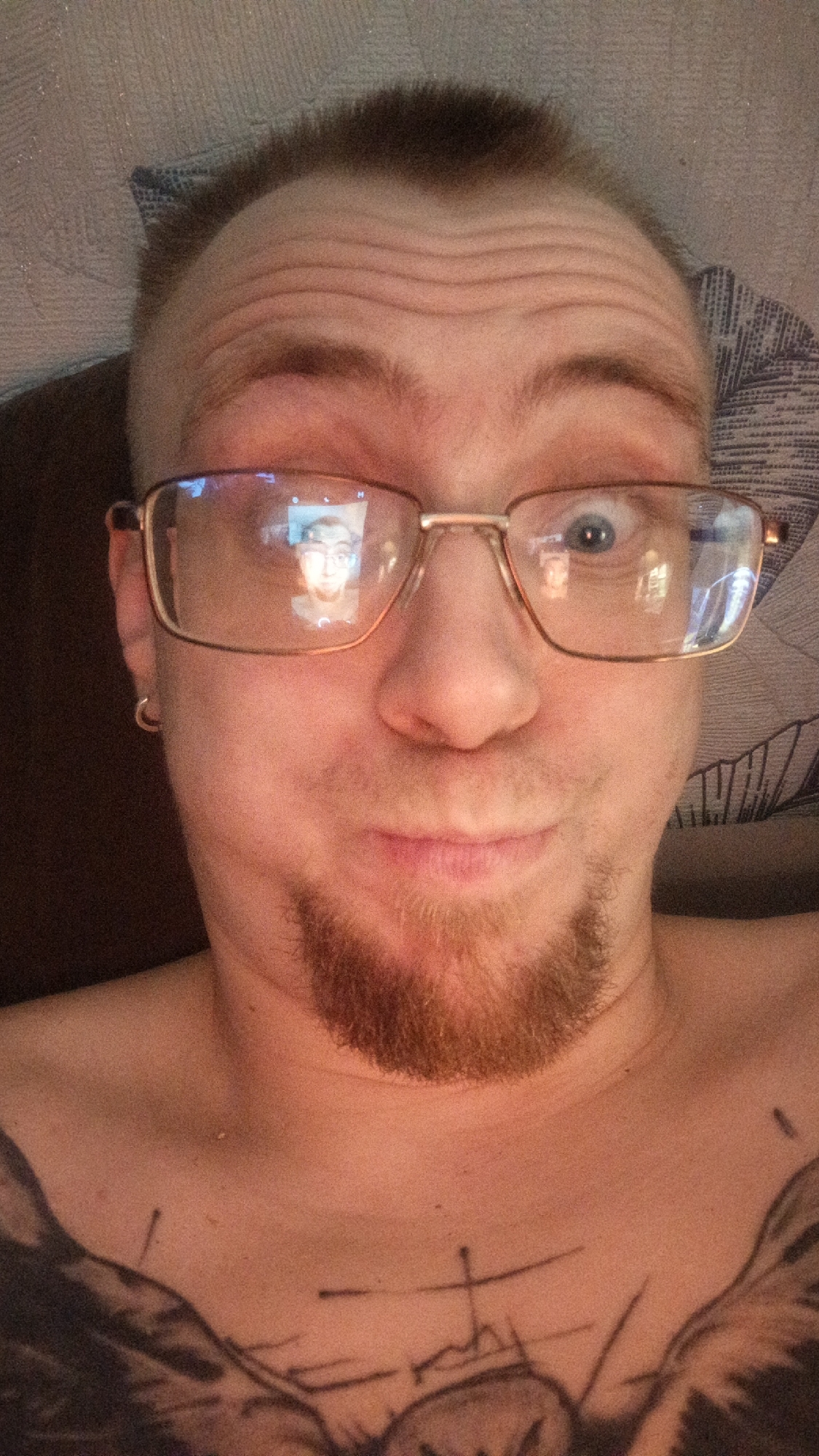 A close-up, head-and-shoulders, selfie-style portrait shows a light-skinned person with fair skin, a light brown goatee, and short dark hair. The individual is wearing gold-rimmed glasses and a hoop earring in their left ear. Their forehead displays prominent horizontal wrinkles, and their right eye is wide open, while the left eye is partially obscured by the glasses. A reflection of a person wearing glasses is visible in the lenses of the individual's glasses. The person's chest is visible, with a tattoo featuring abstract lines and shapes, including what appears to be a cross. The background is a textured, light-colored surface, possibly a wall or bedding. The lighting is warm and appears to originate from the front.