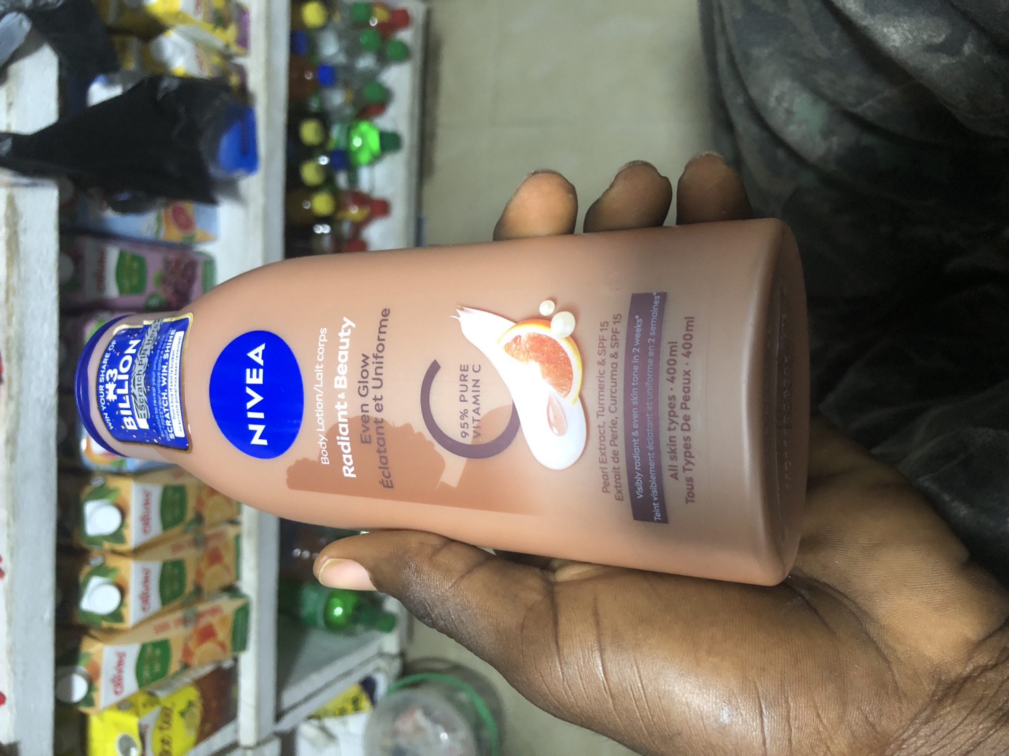 A person's hand holds a Nivea Radiant & Beauty Even Glow body lotion bottle in front of a shelf filled with various products, including juice boxes and bottled drinks. The bottle is pink and features a blue Nivea logo and text detailing the product's benefits and ingredients. The background suggests a retail setting, possibly a small shop or market stall in Ado, Nigeria. The lighting is artificial and somewhat dim, casting shadows on the objects.