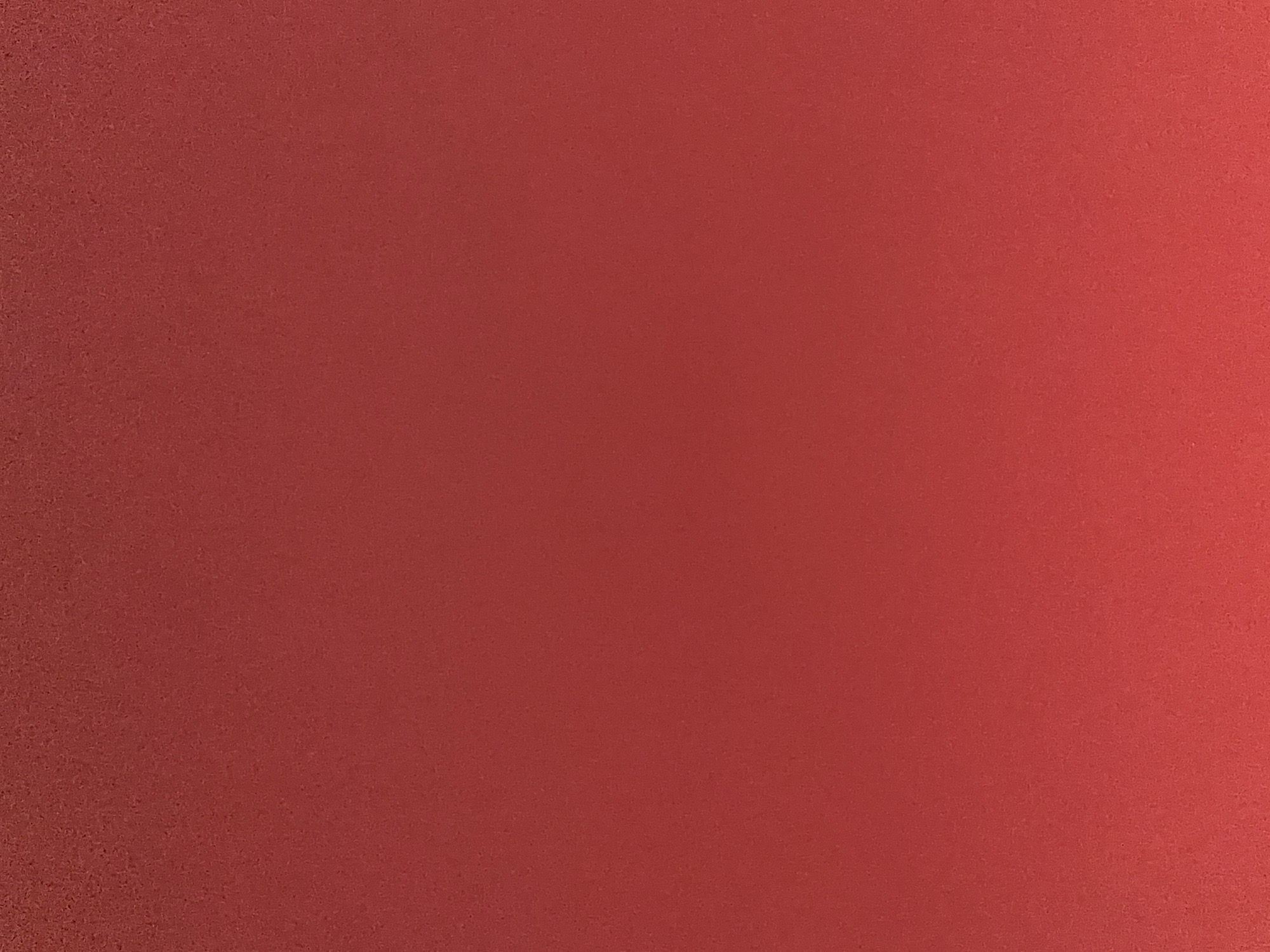 This media file is a still image of a textured, reddish-orange surface. The surface appears to be a wall or a similar material, with a fine grain visible throughout. There is a subtle gradient of color, with the left side being slightly darker and more textured, and the right side becoming lighter and smoother, suggesting a light source from the right. No people, objects, scenes, activities, or text are present. The image is abstract and focuses solely on the texture and color of the surface.