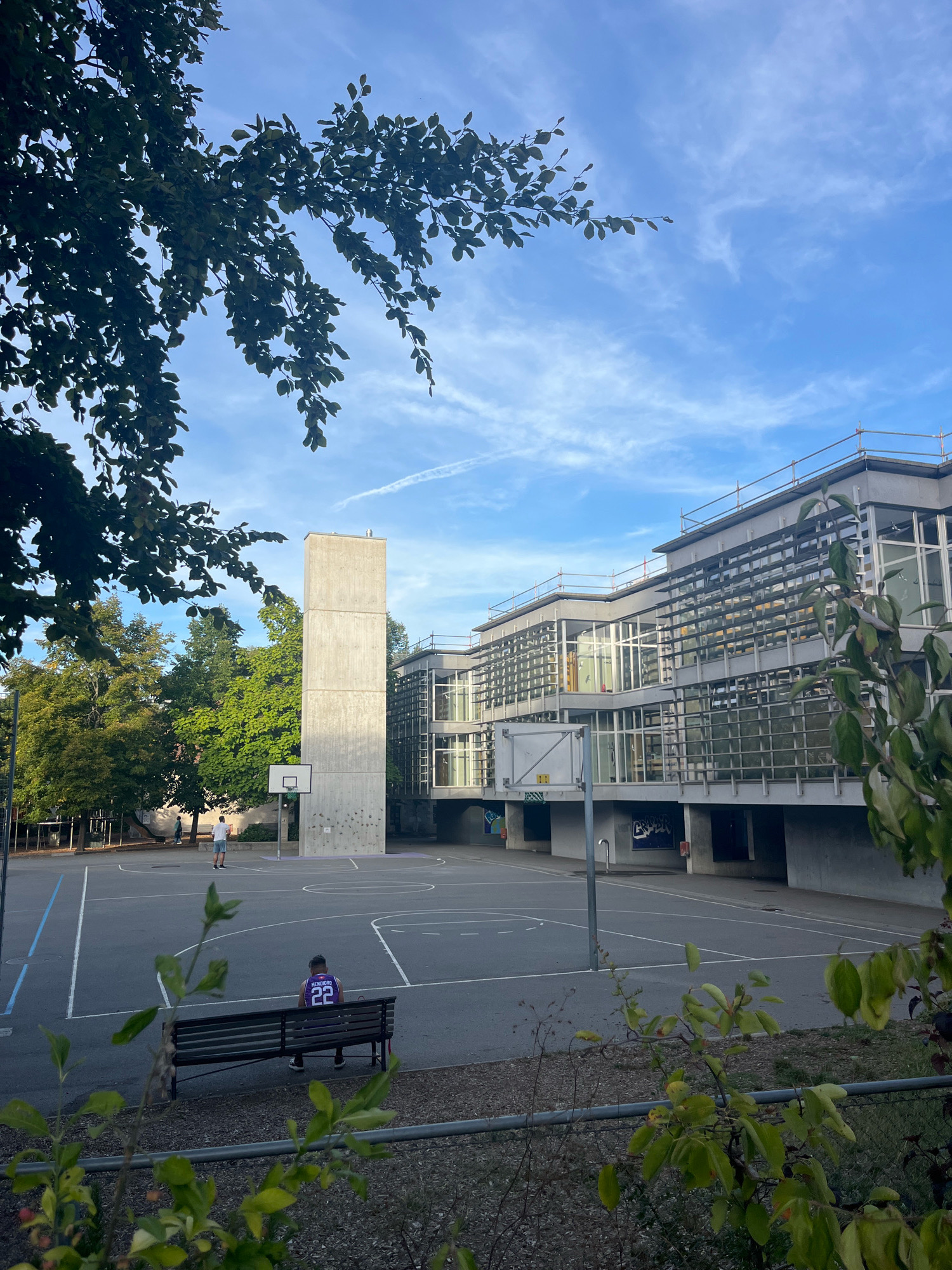 This is an exterior, eye-level, daylight, full shot of a schoolyard with a basketball court in Geneva, Switzerland. In the foreground, a person is sitting on a bench with their back to the camera. They are wearing a purple basketball jersey with the number 22 on it. A second person is standing on the basketball court, facing away from the camera. Behind them, a tall concrete structure stands next to a modern building with many windows and scaffolding. Trees with green foliage surround the area. The sky is blue with wispy clouds.