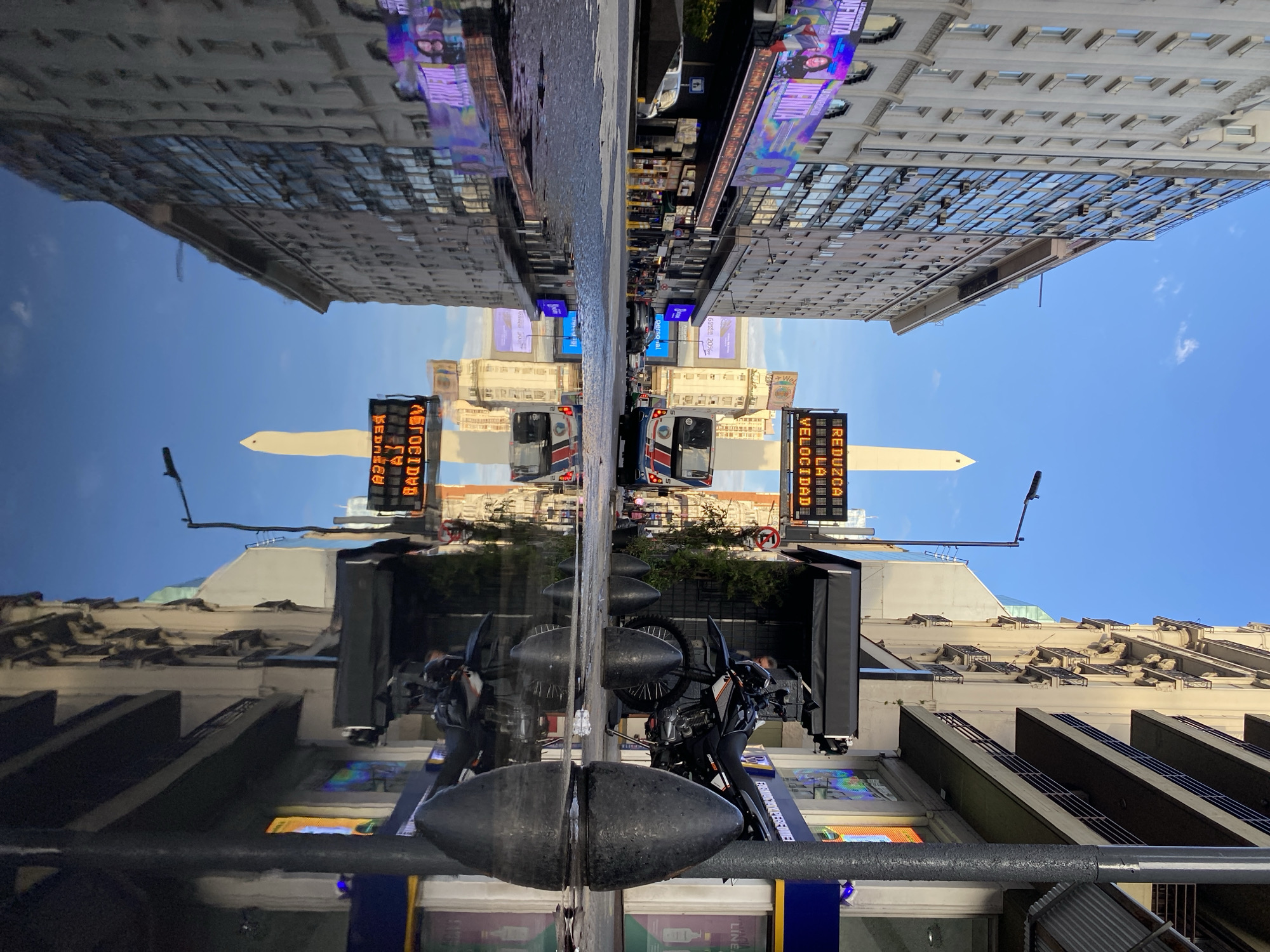 The image shows a reflection of a street scene in Buenos Aires, Argentina. The reflection seems to be from a wet surface or a glass panel. The Obelisco monument is visible in the background, along with buildings lining the street. There is a motorcycle parked in the foreground, and various digital billboards and traffic signs are visible. There are buses or similar vehicles in the middle distance.