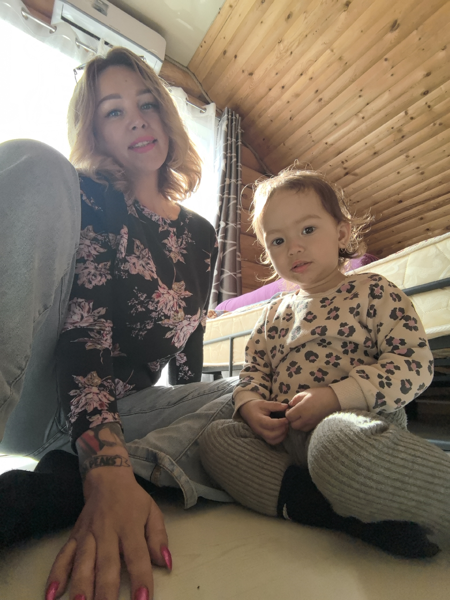 A woman and a young child are sitting on the floor in an indoor space with a wood-paneled ceiling. The woman is wearing a floral print shirt and light-colored jeans, and has a tattoo on her forearm. The child is wearing a patterned sweatshirt and gray pants. A bed is visible in the background, along with curtains. The specific location is not discernible as being within Noviy, Russia, based on visual elements alone.