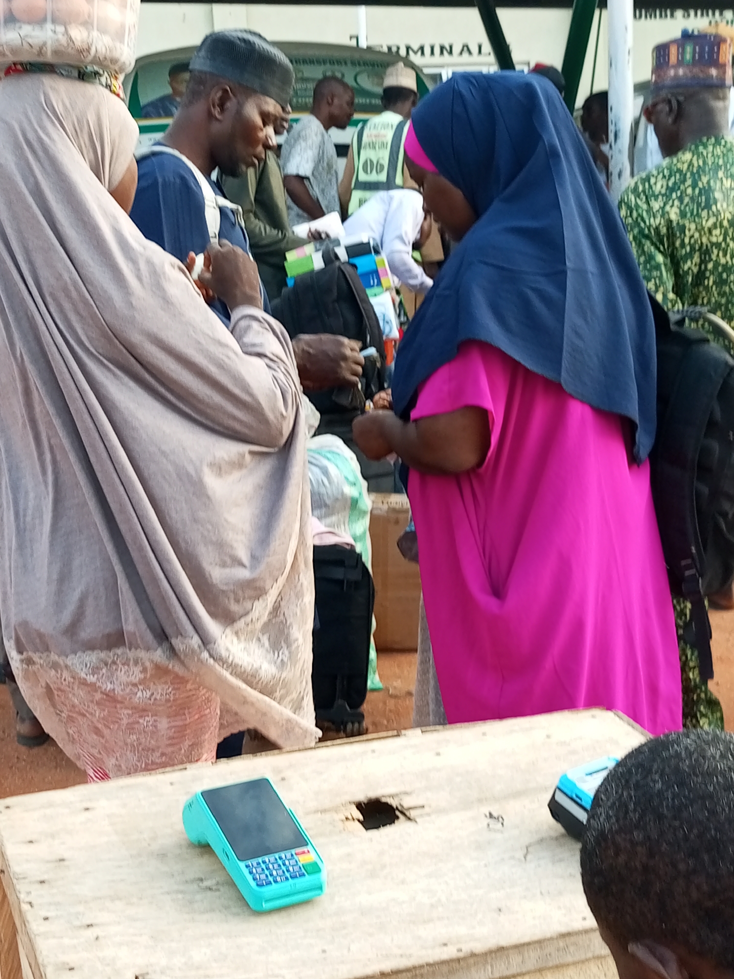In Gombe, Nigeria, a group of people are gathered around a transaction. In the foreground, a teal-colored point-of-sale device rests on a wooden surface. A woman wearing a hijab and a vibrant pink dress stands behind the device, her hands near it. To her left, another woman, also in a hijab, is positioned with her back to the camera, carrying a basket of items on her head. A man wearing a blue garment and a cap is visible to her side, facing away. In the background, other individuals are present, some wearing vests with numbering, suggesting an organized setting like a transport terminal. The environment appears to be outdoors, with hints of a vehicle and signage in the distance, indicating it is likely a busy public space.
