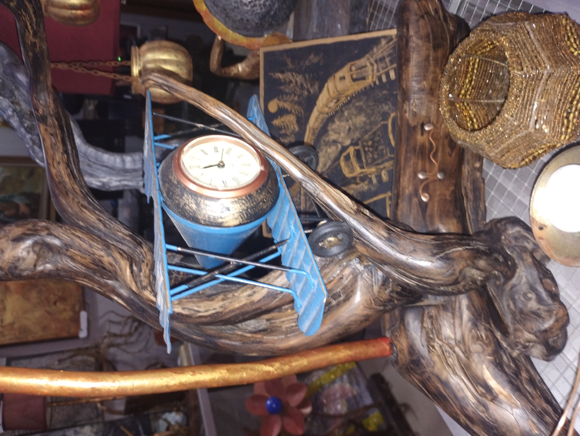 This is a close-up, medium-angle, indoor shot of a collection of decorative objects. The primary subject is a rustic, artistic arrangement featuring elements that resemble driftwood, possibly accented with metallic paint or finishes, creating a dark, aged, and textured appearance.
Prominently featured within the arrangement is a vintage-style clock, its face white with Roman numerals and black hands. The clock is housed in a dark, possibly metallic casing, with a copper-colored rim. It is suspended within a blue, airplane-shaped structure, suggesting a whimsical or artistic representation of flight. The airplane has blue wings and a cockpit area, with visible black wheels.
Other objects in the scene include a gold-colored, chain-like element that appears to be suspended. To the right of the clock and airplane, there is a spherical, decorative item covered in small, shiny beads, predominantly gold and amber in color. Further to the right, a bright white, circular object is visible, possibly a light source or another decorative element. Behind the clock and airplane, a dark panel with gold or yellow intricate designs is partially visible. Below the main arrangement, a long, golden-colored rod or pipe runs horizontally across the frame. At the bottom center, a small, stylized flower with dark red petals and a blue center is visible.
The lighting appears to be artificial, casting soft shadows and highlighting the textures of the objects. The overall impression is one of an eclectic display of decorative items, possibly in a home or a shop specializing in rustic or artistic decor. There is no visible text in the image. The scene does not depict any people or specific weather conditions.